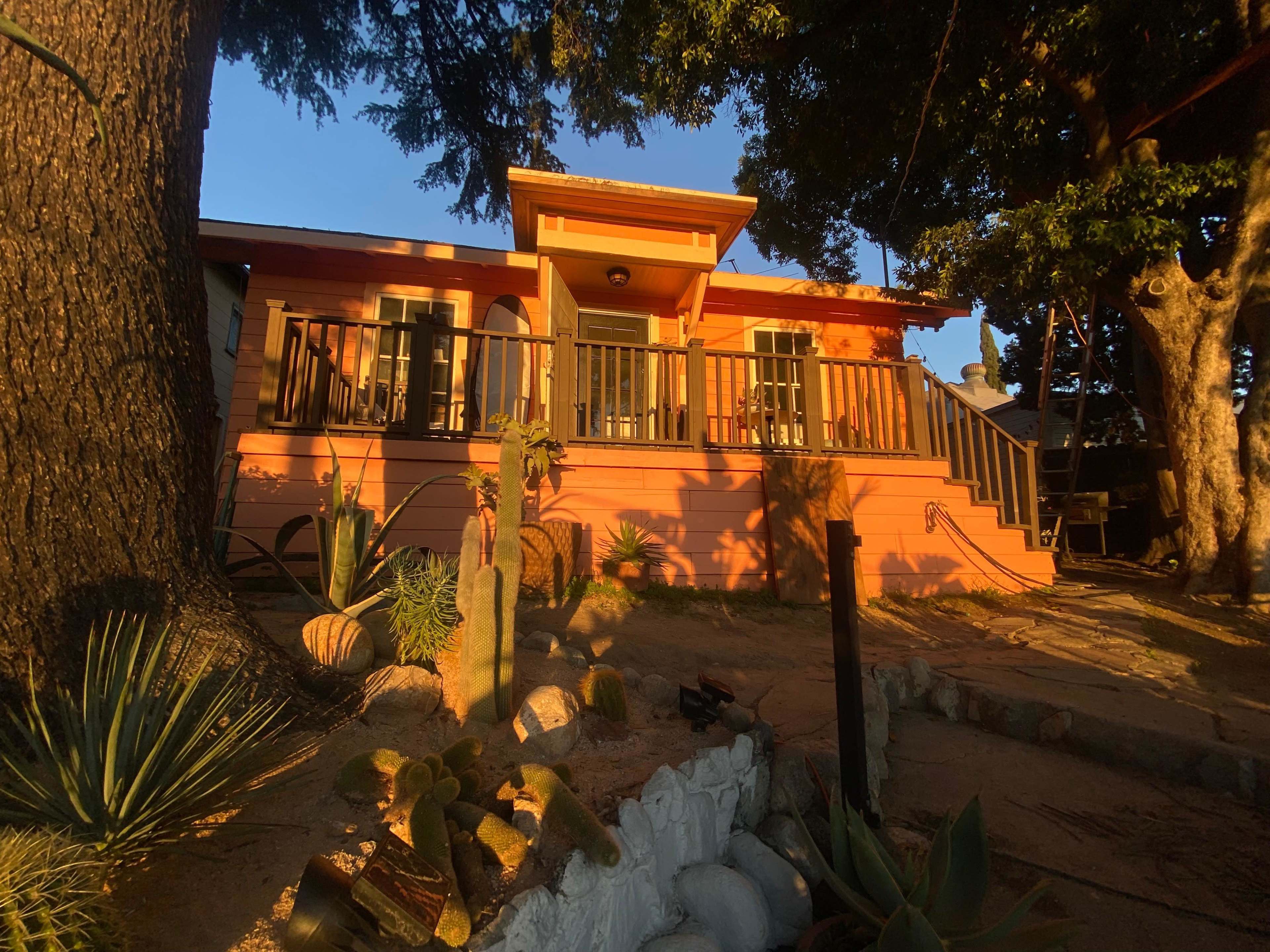 A colorful house with a deck and chairs is set among trees and various plants in a garden at sunset.