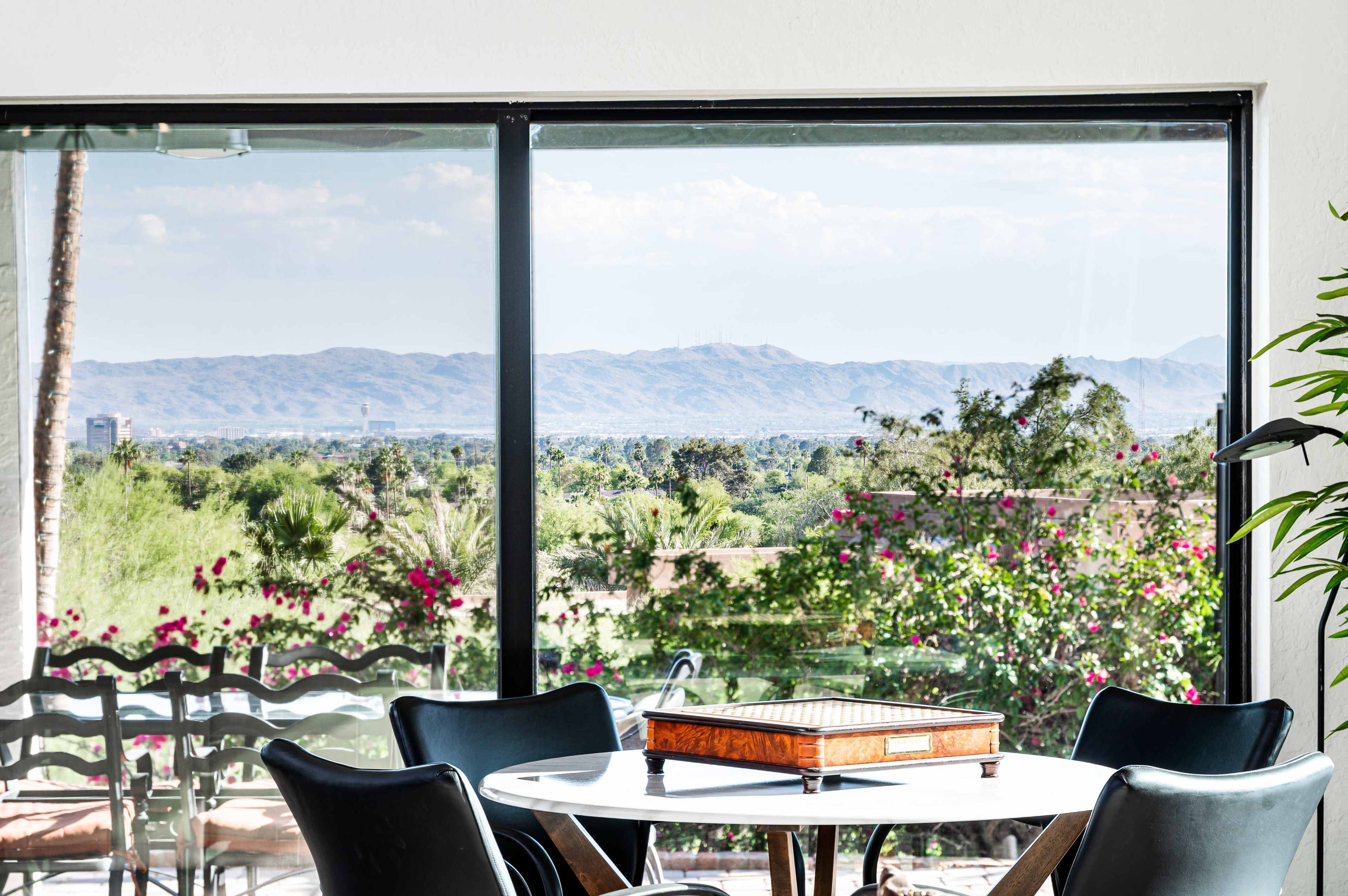 A modern dining area features a round table with a game board, overlooking flowering plants and distant mountains through large windows.