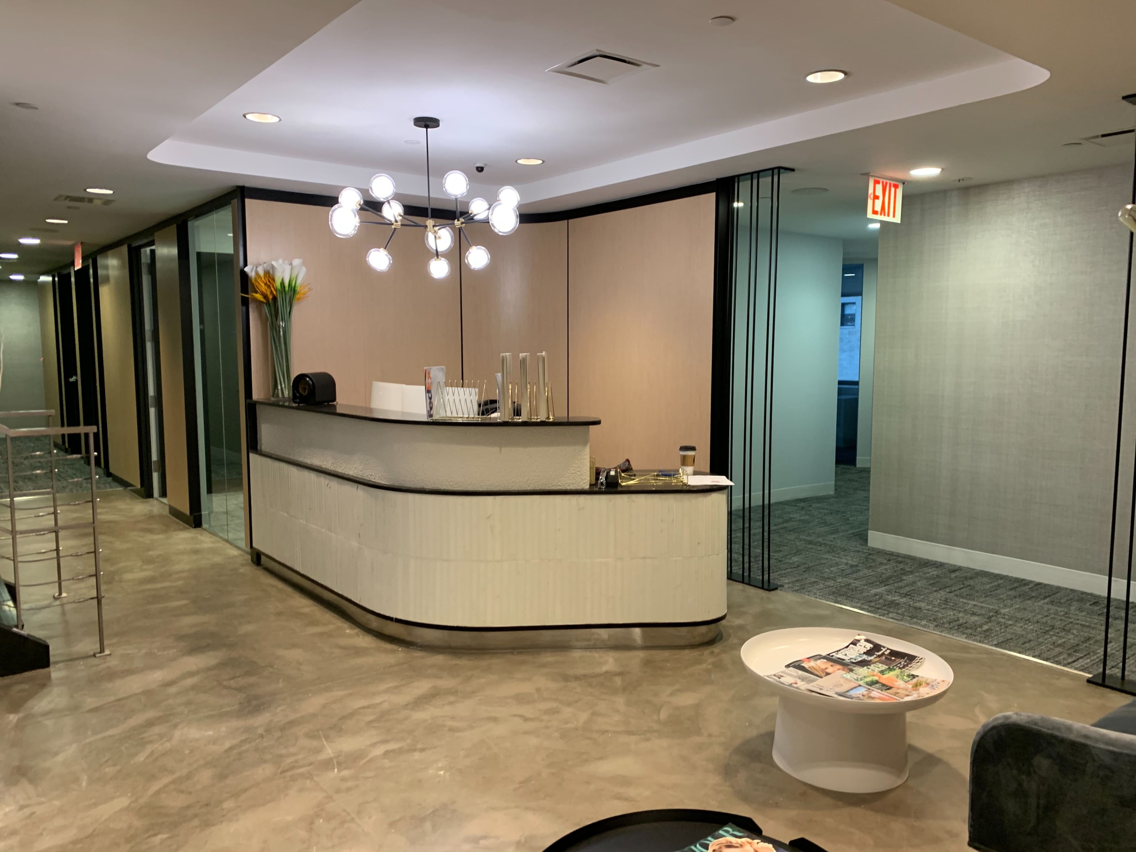 The image shows a modern reception area with a curved front desk, a decorative light fixture, and glass office partitions.