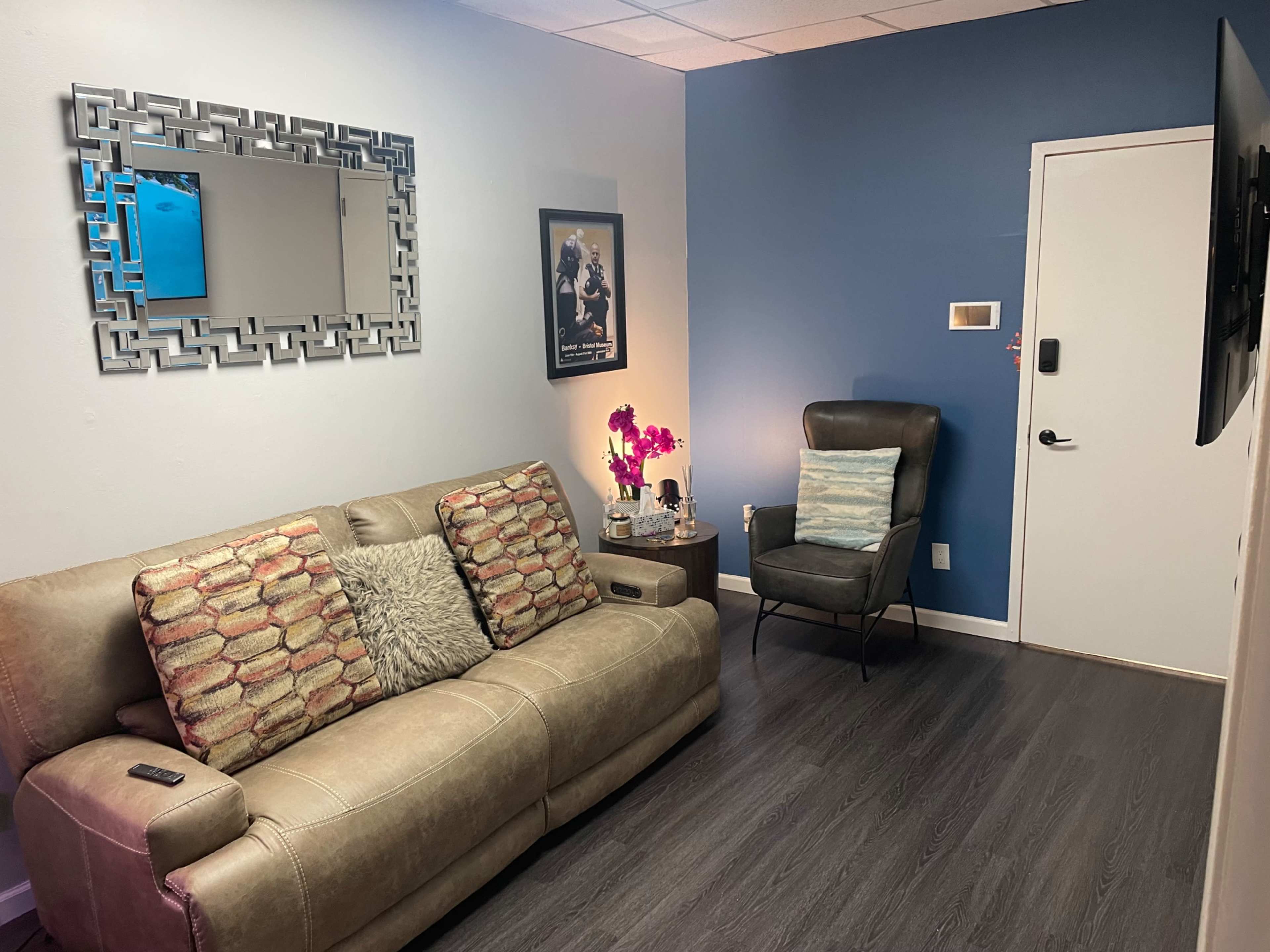 A small living room features a beige couch with decorative cushions, a gray armchair, a wall-mounted TV, and a blue accent wall.