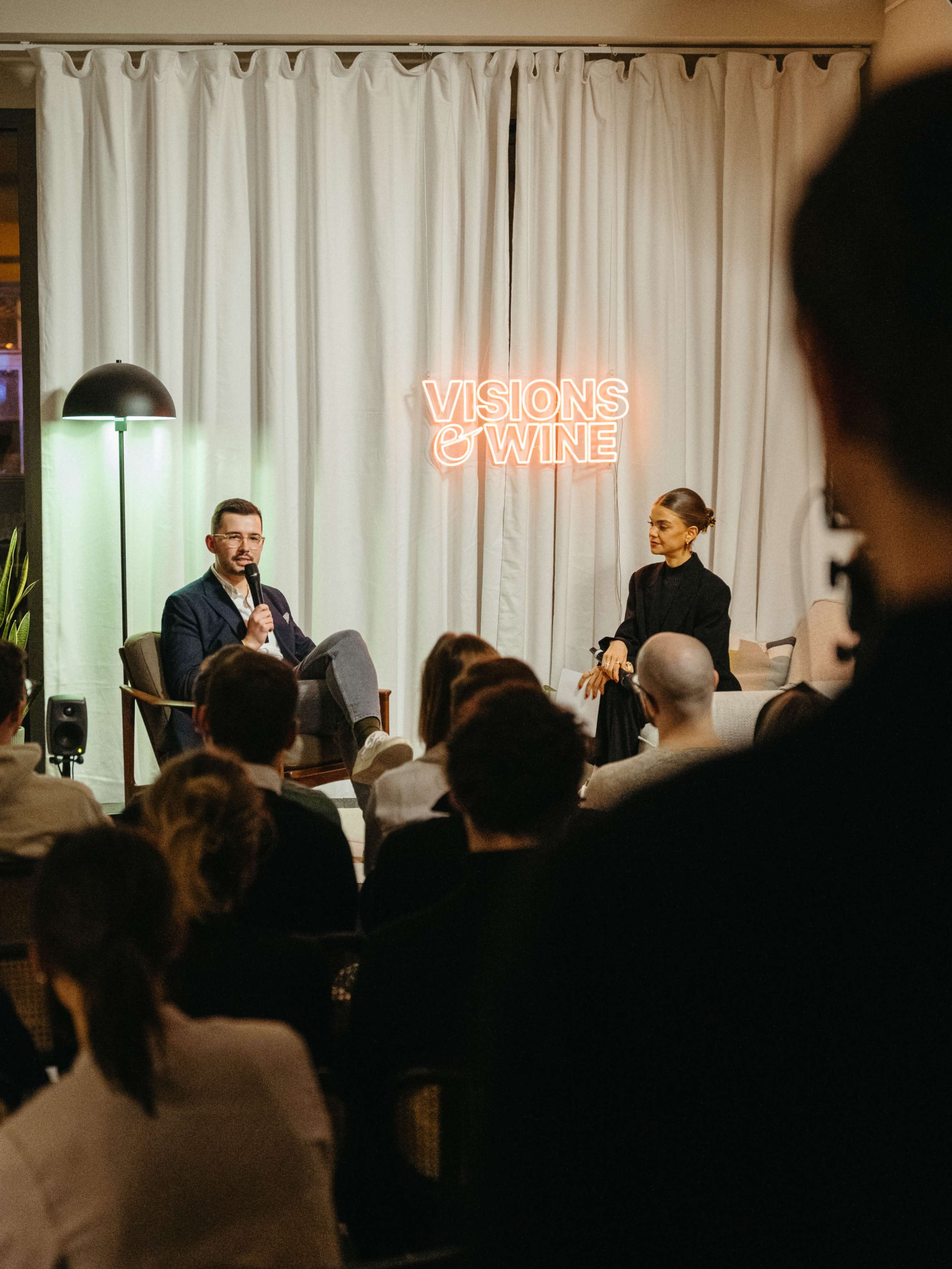 A man and a woman sit in front of an audience, discussing topics related to wine, with a neon sign reading "VISIONS OF WINE" in the background.