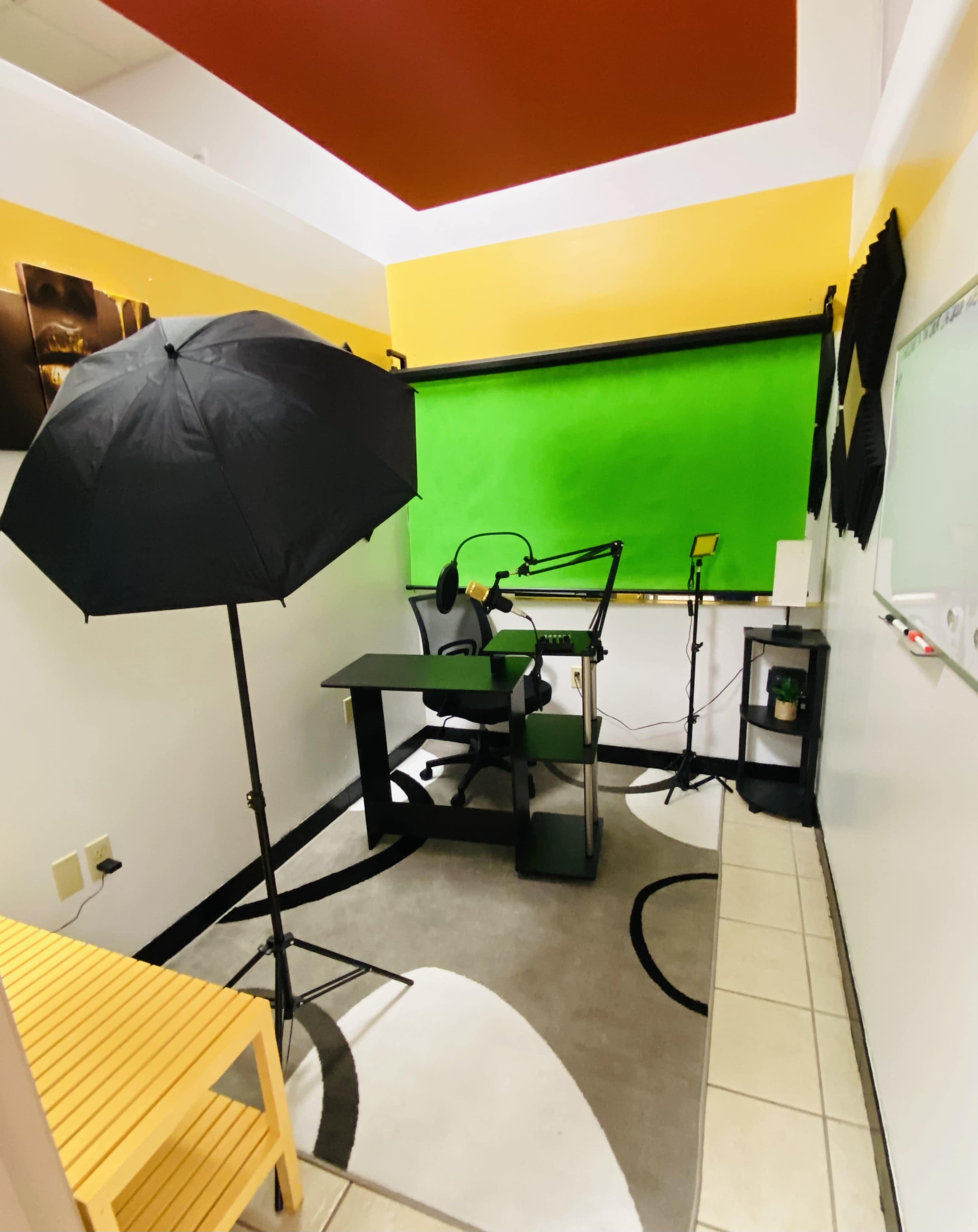 A small recording studio with a green backdrop, a microphone setup, a black umbrella light, a desk, a chair, and a whiteboard on the wall.