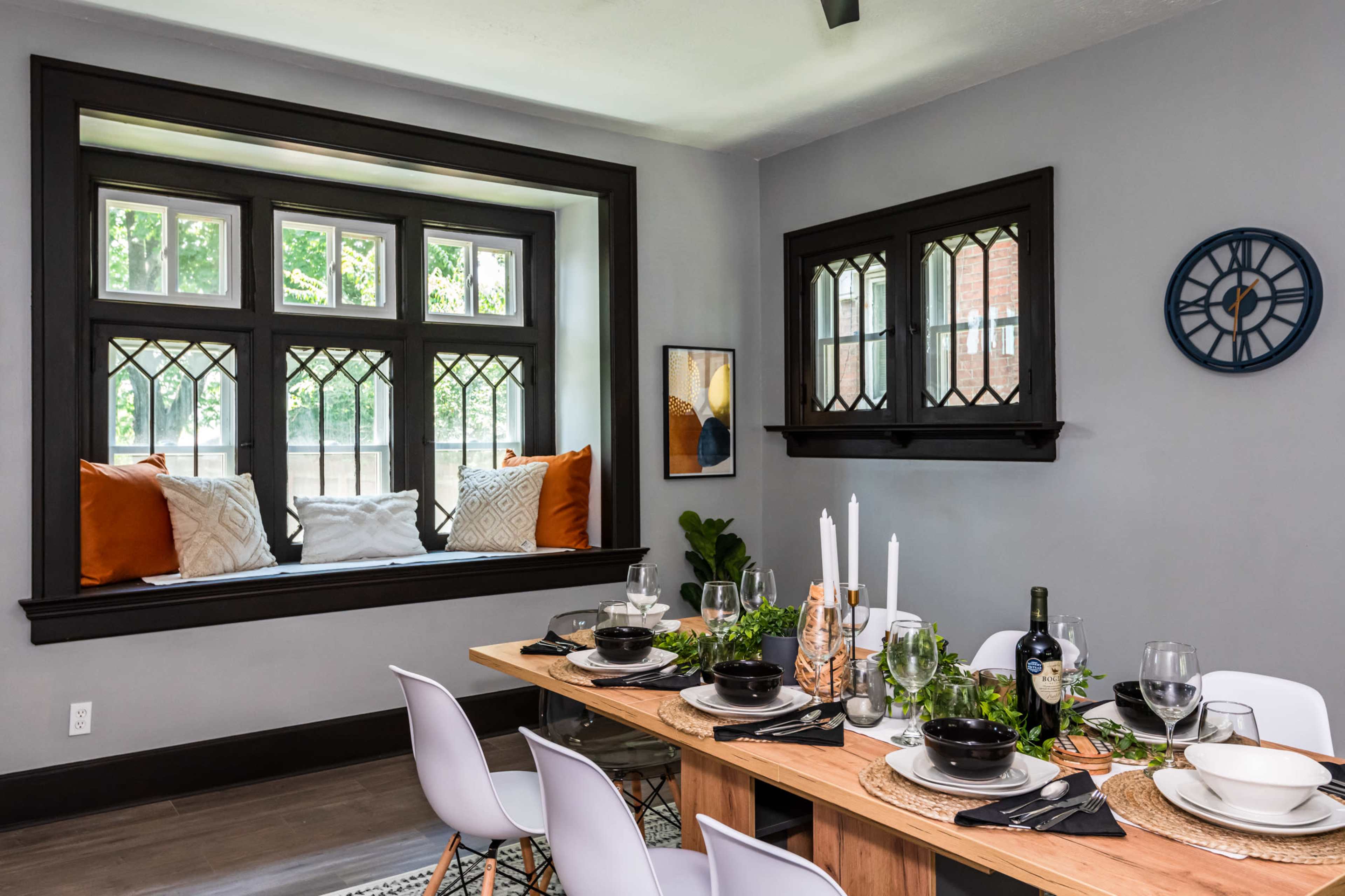 A dining area features a long wooden table set with black dishes, wine glasses, and candles, alongside a cozy window seat adorned with decorative pillows.