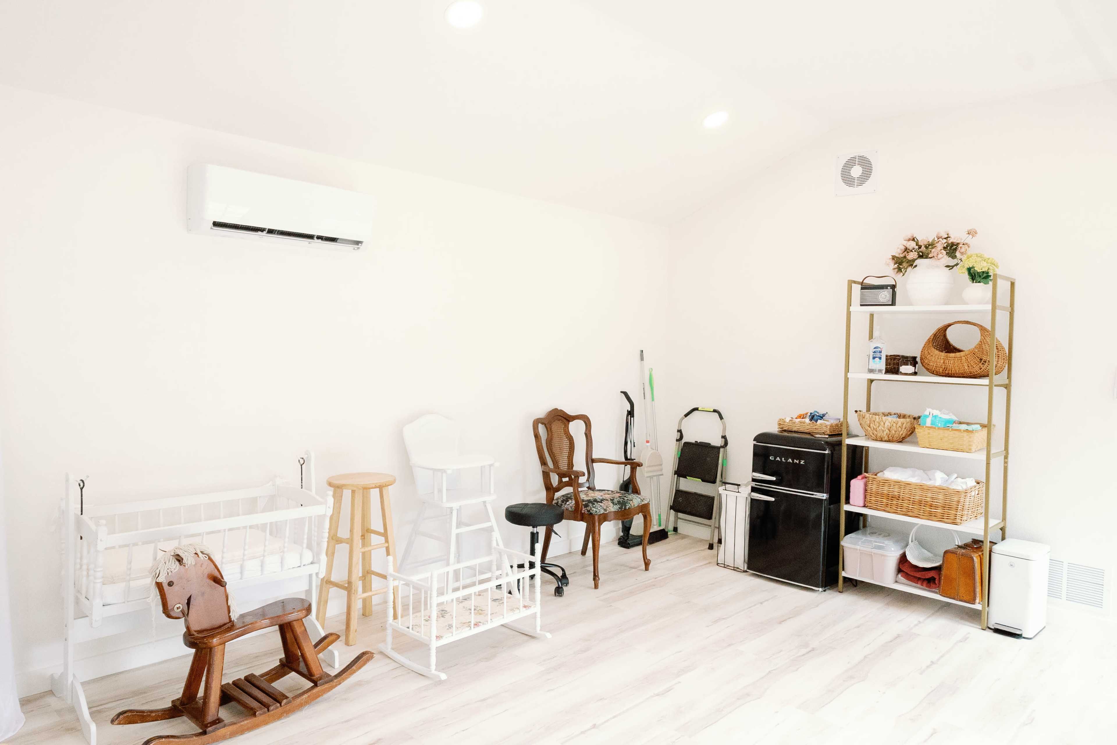 The image shows a spacious, well-lit room furnished with a rocking horse, a high chair, a folding step stool, and a small black refrigerator next to a shelving unit filled with baskets and storage containers.
