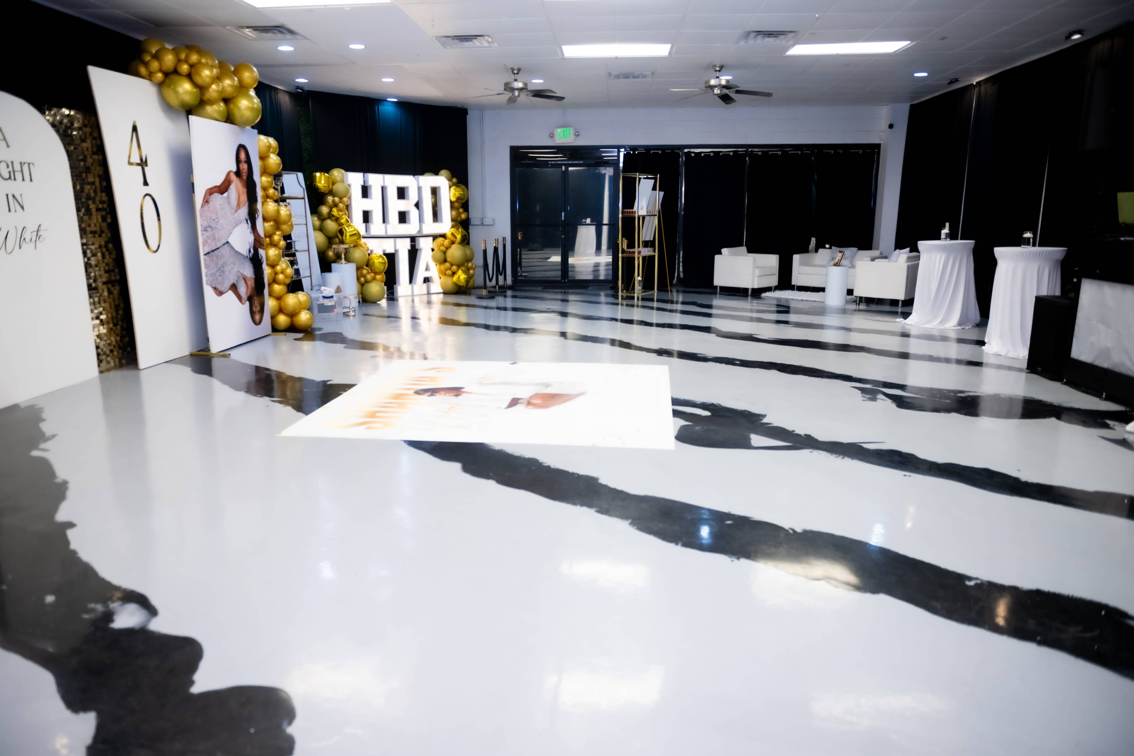 The image shows a decorated event space featuring a white and gold balloon arch, a large "HBD" sign, and a striped floor design, set up for a celebration.
