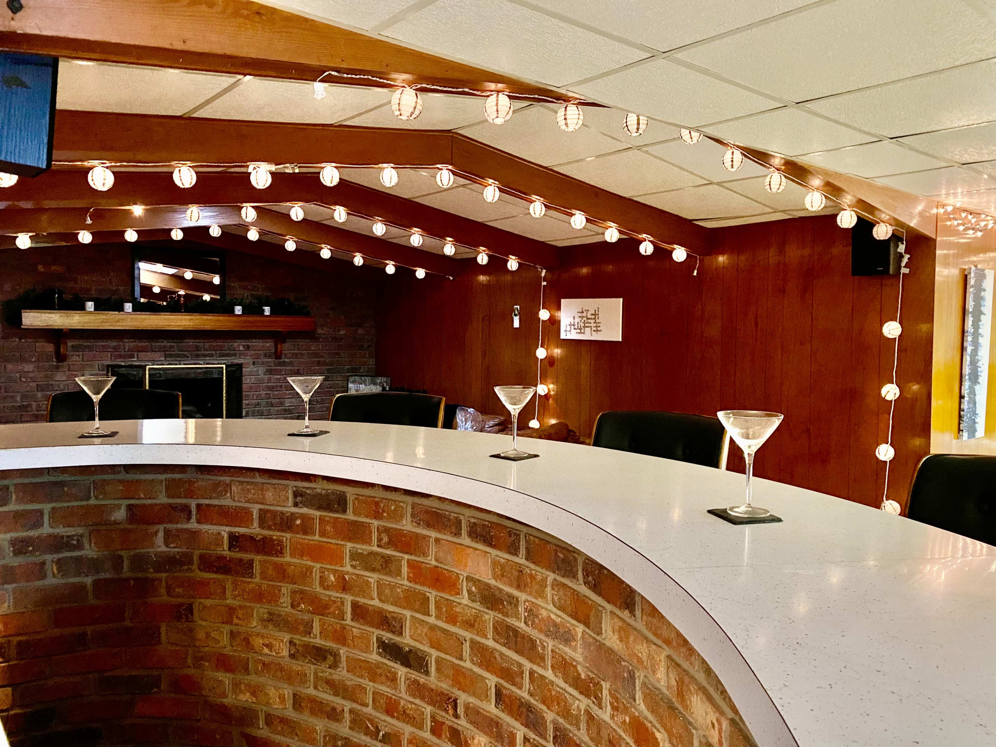 A curved bar with martini glasses sits under string lights in a cozy room featuring wooden paneling and a brick wall.