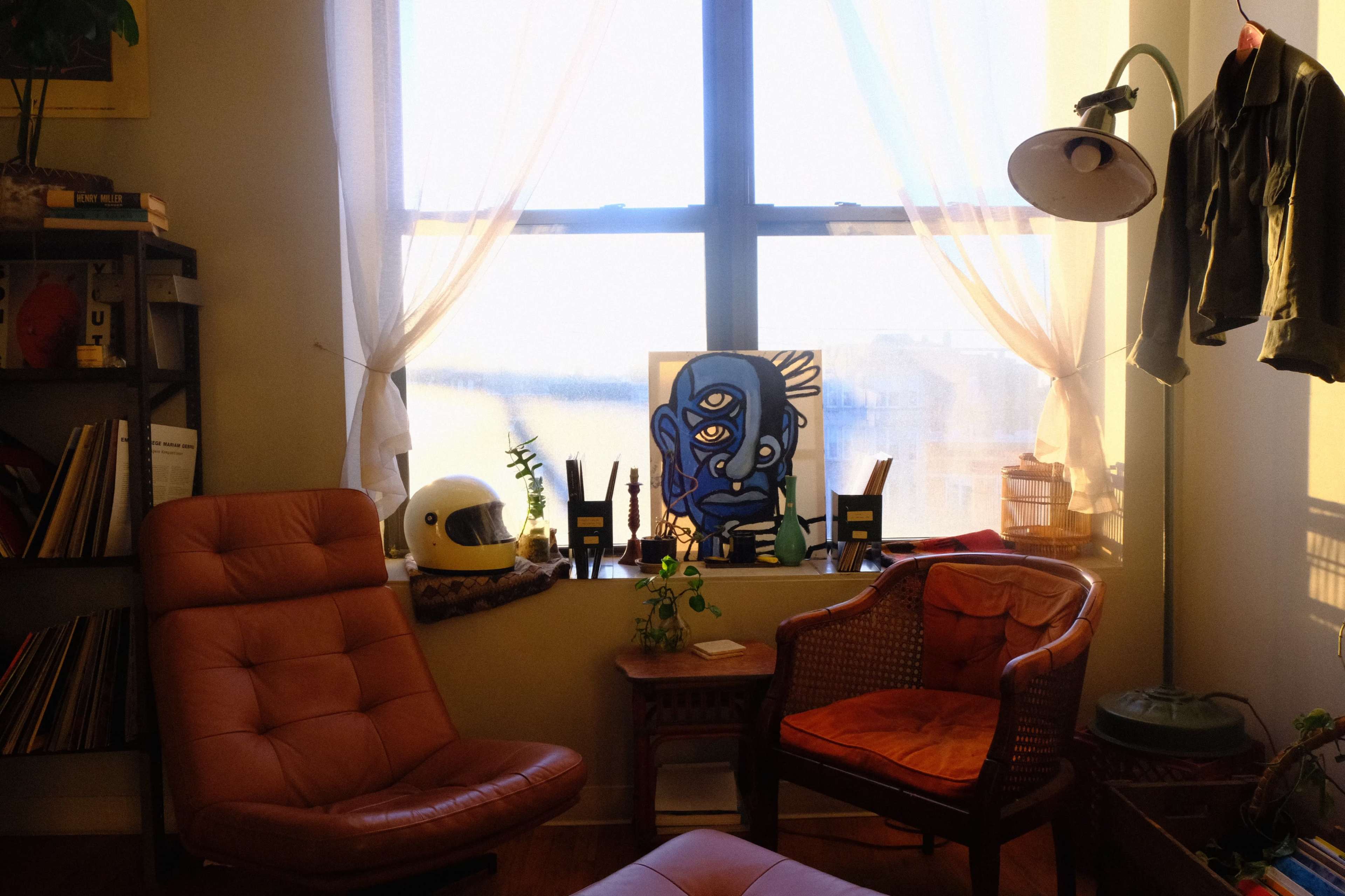 The image shows a cozy room with two chairs, a potted plant, and a colorful painting on a windowsill illuminated by natural light.