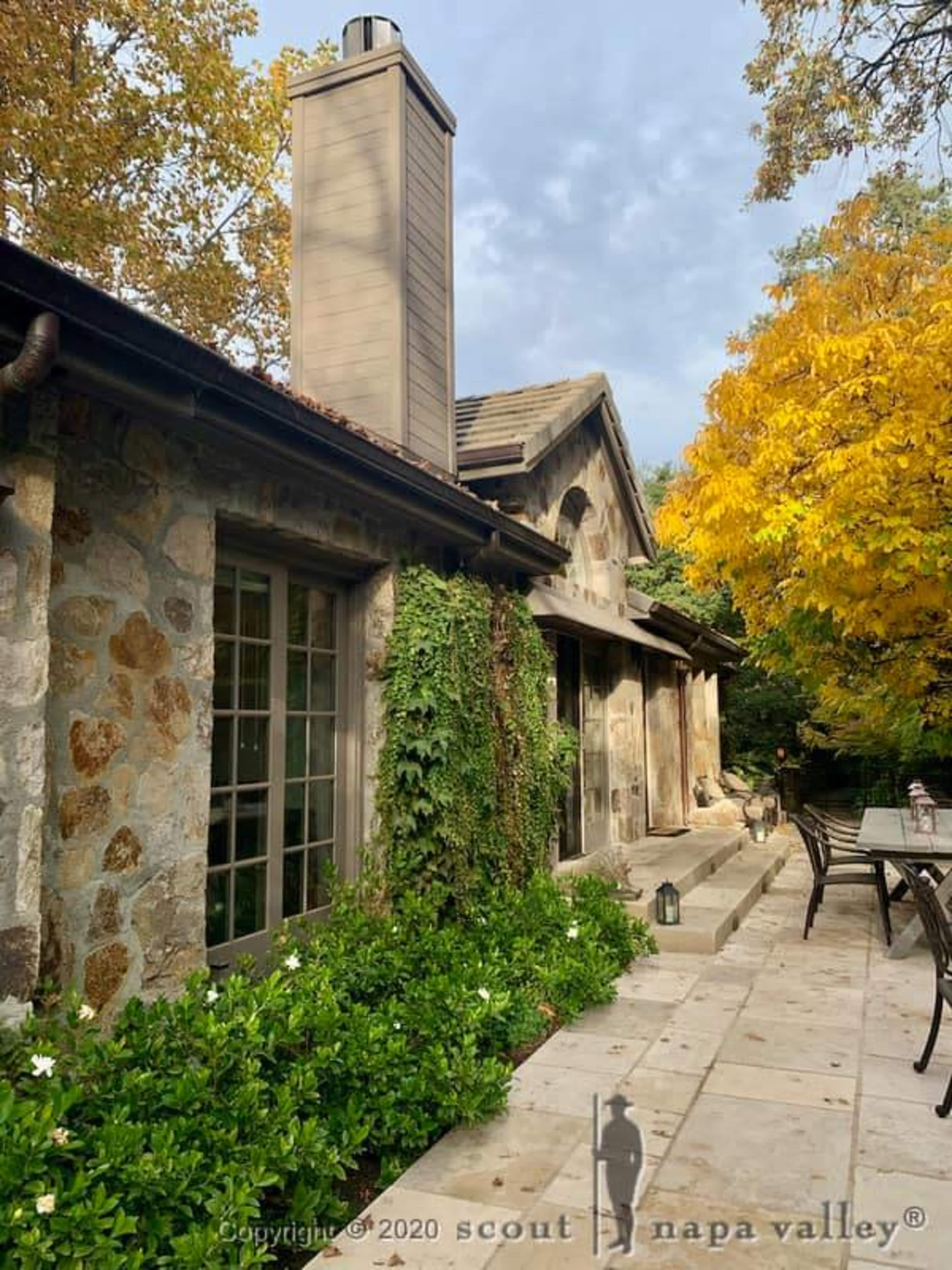 A stone and wood house with a chimney, surrounded by green foliage and a tree with yellow leaves.