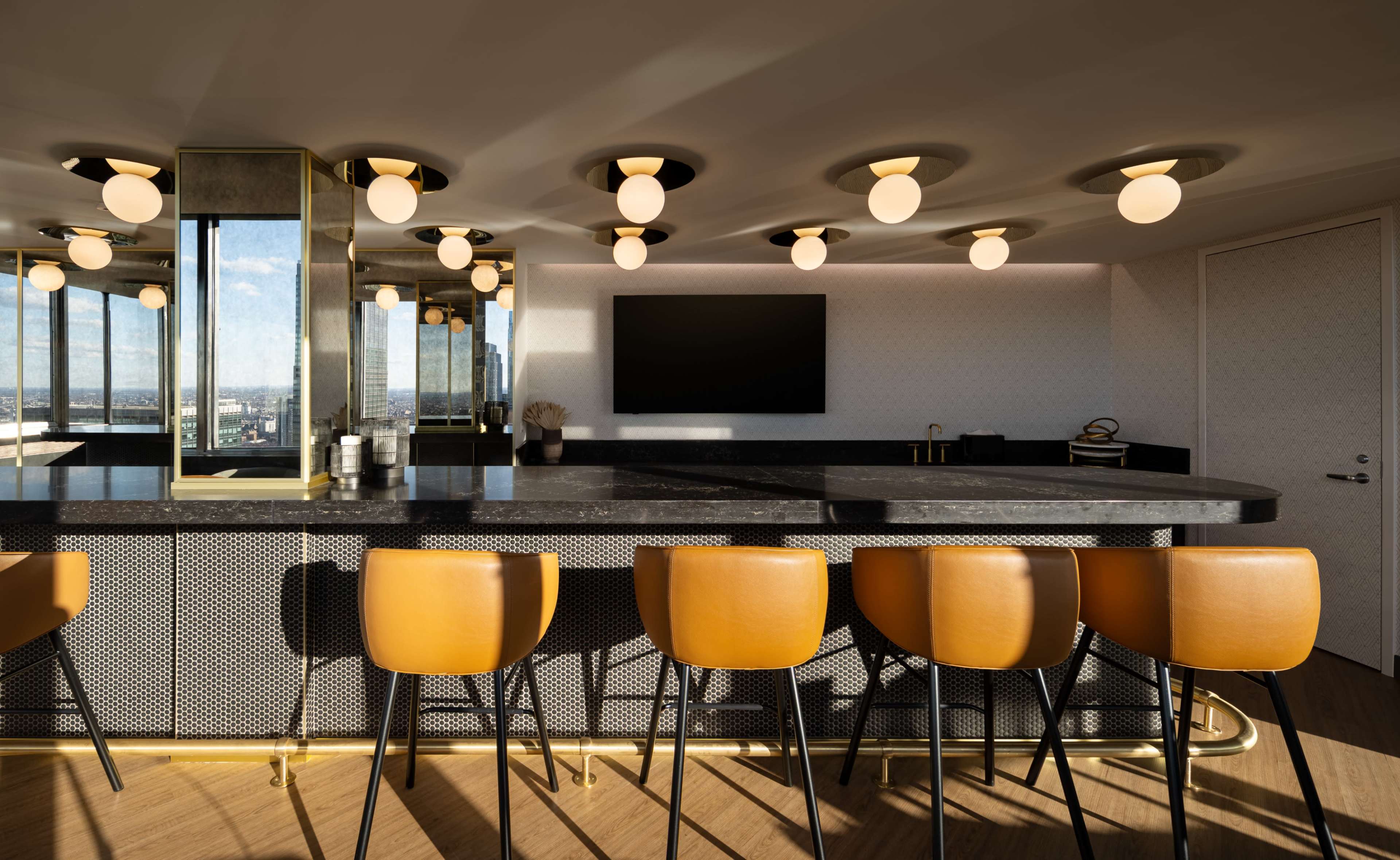 A modern bar area features a sleek, dark countertop, golden lighting fixtures, and orange stools arranged in front.