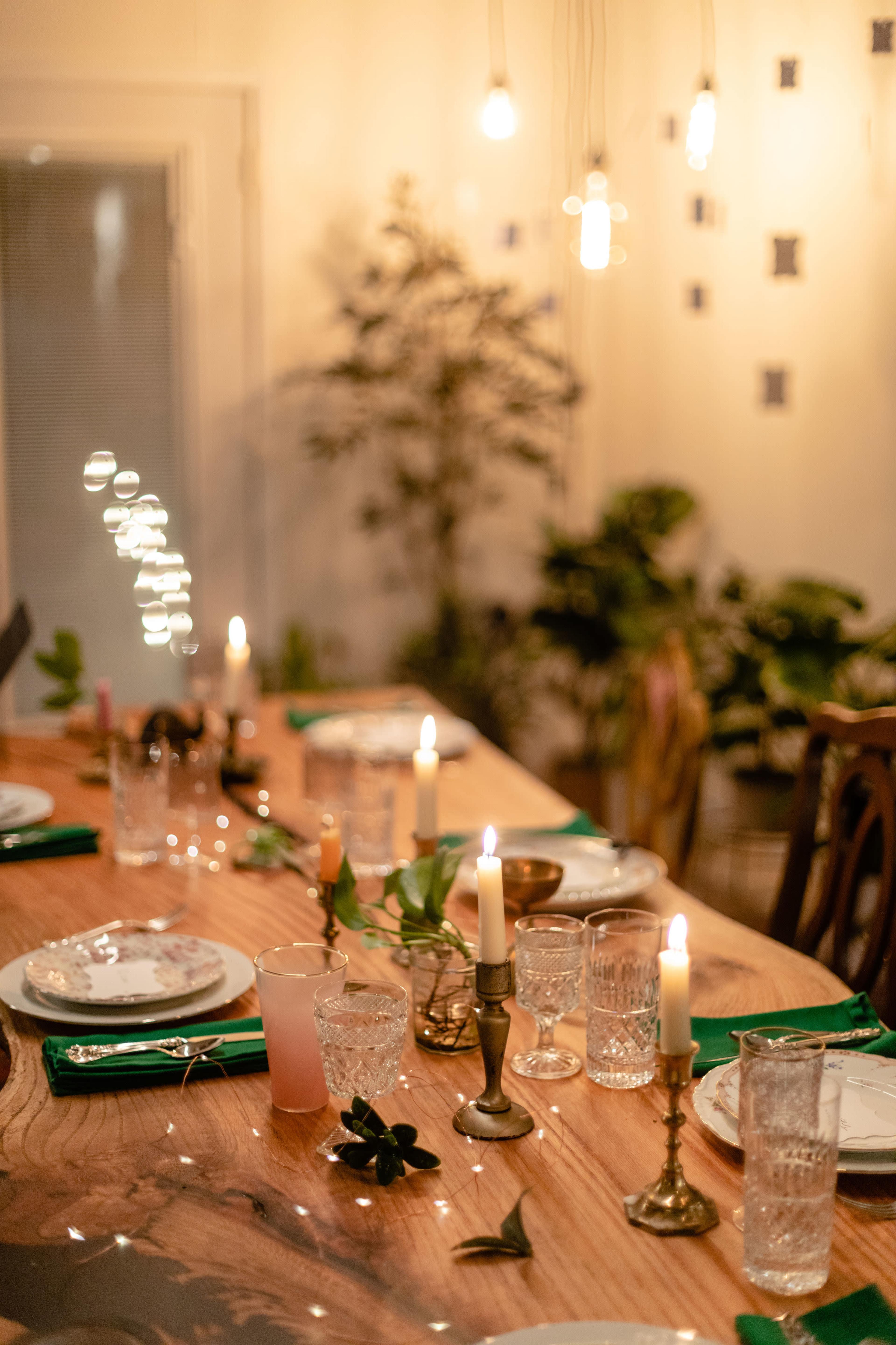 A dining table is elegantly set with green napkins, glassware, and candles, surrounded by plants and illuminated by hanging lights.