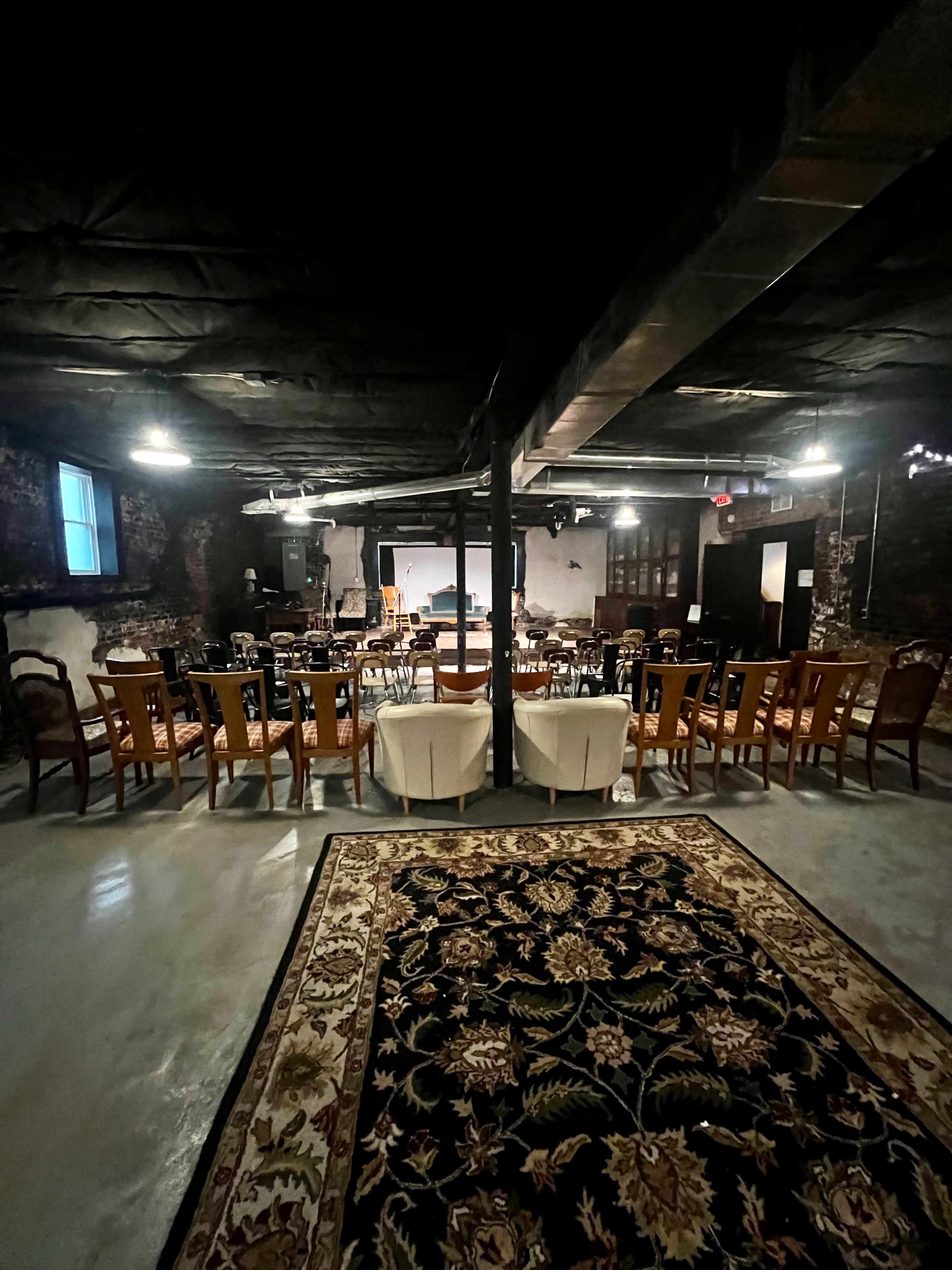 The image shows a dimly lit basement venue set up with rows of chairs and a central stage area, featuring a patterned rug on the floor.