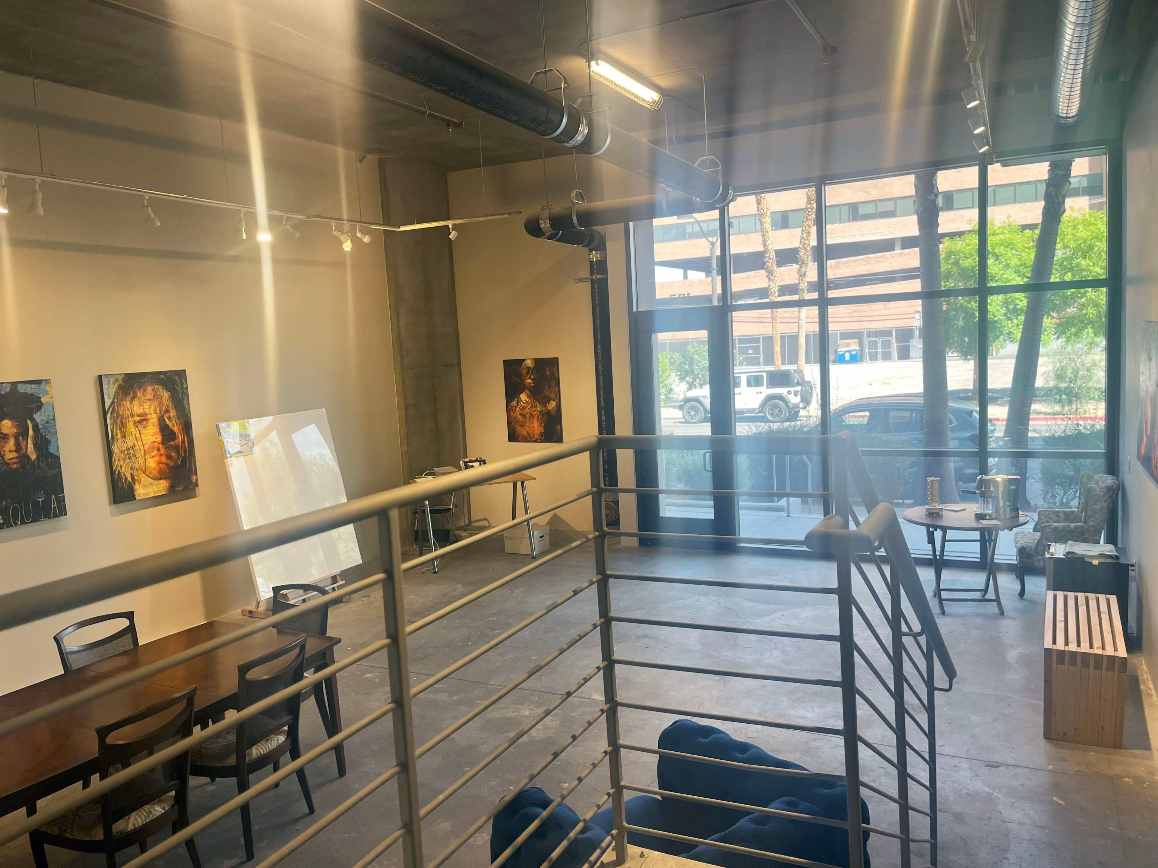 The image shows a spacious art gallery with large windows, featuring several artworks on the walls, a dining table, and a staircase leading to an upper level.