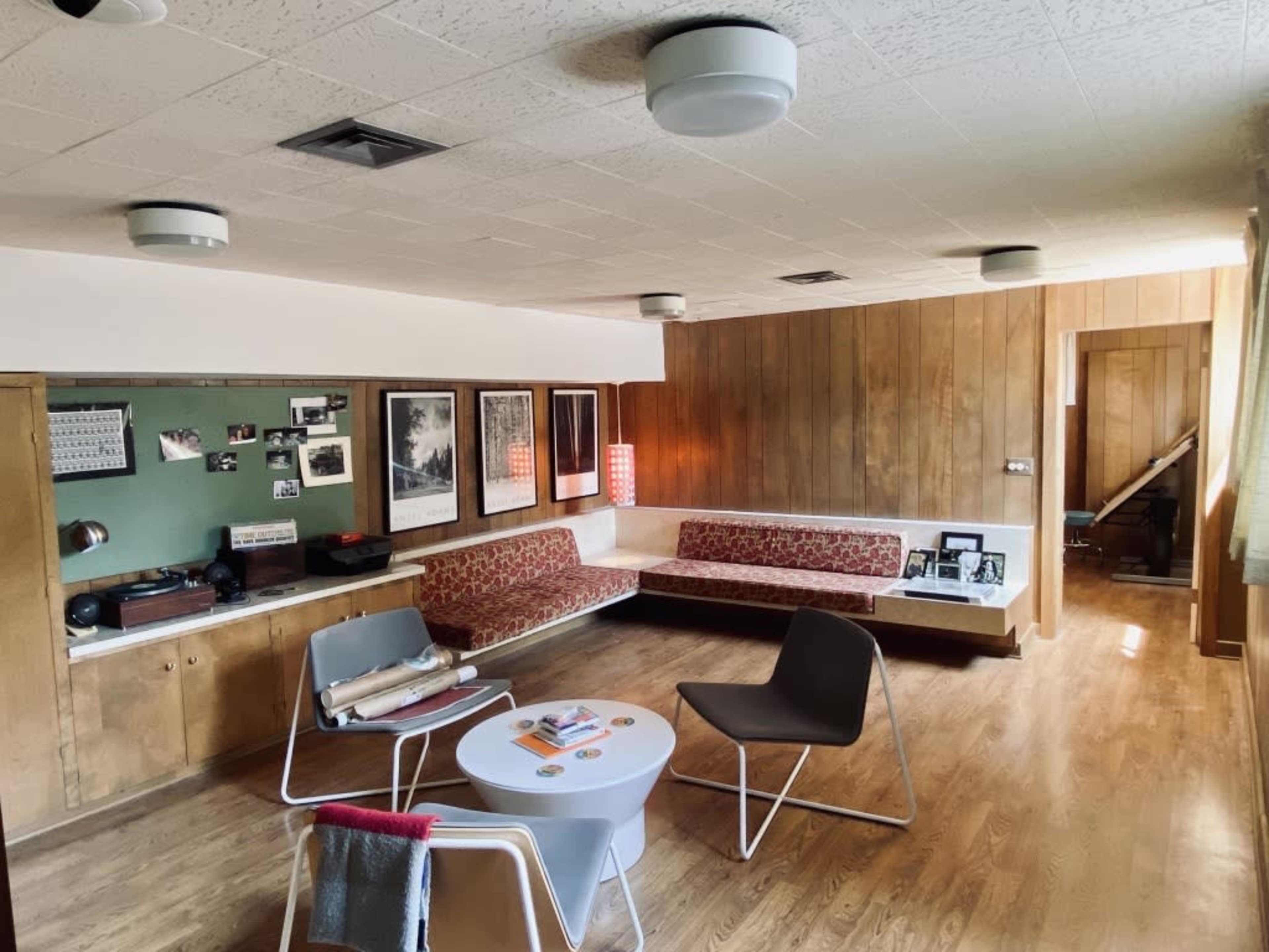 The image shows a mid-century modern living room with wooden paneling, a curved sofa, a circular coffee table, and various framed photographs on the walls.
