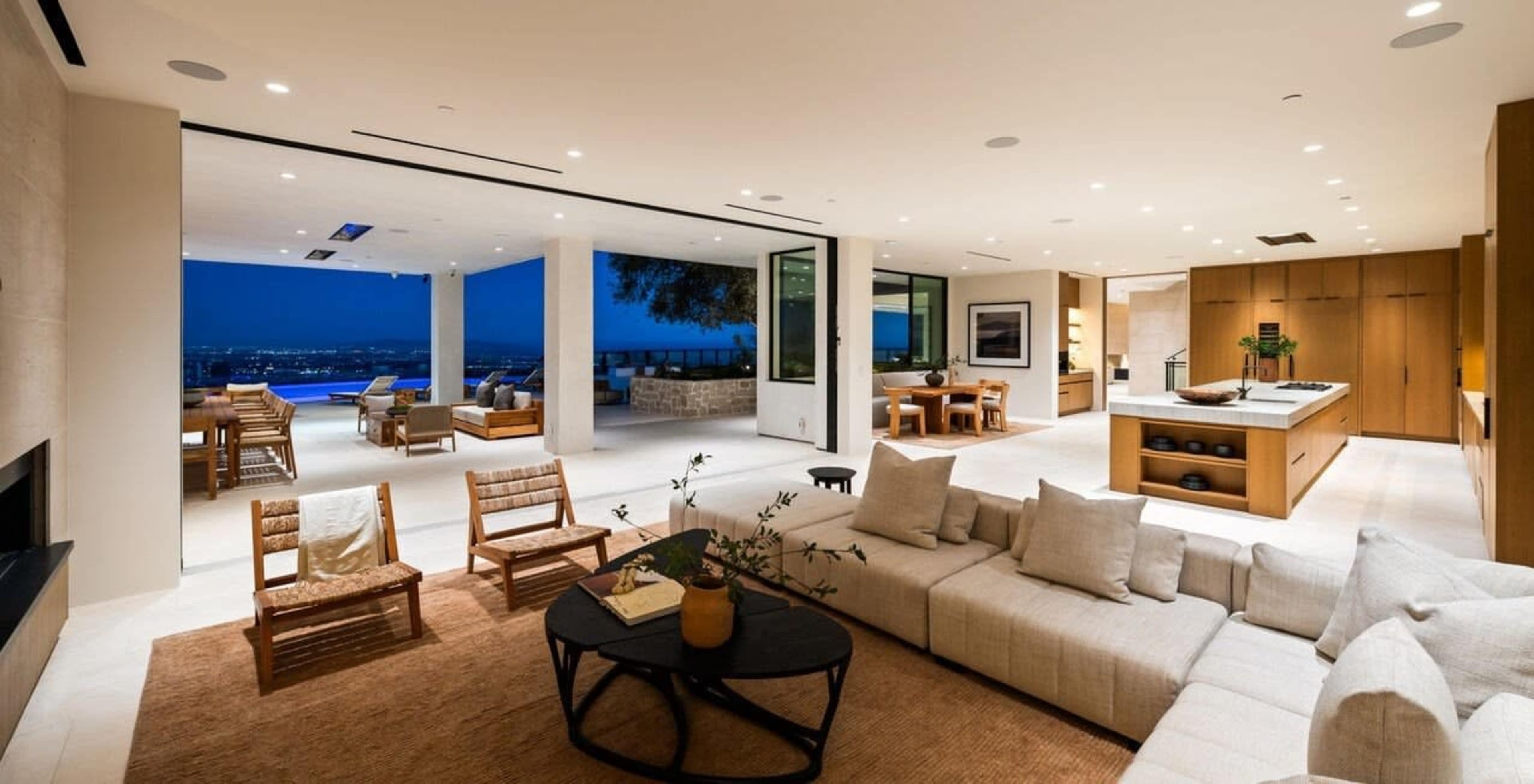 The image shows a modern living space with an open floor plan, featuring a sectional sofa, a coffee table, and a view of an outdoor patio and pool area.