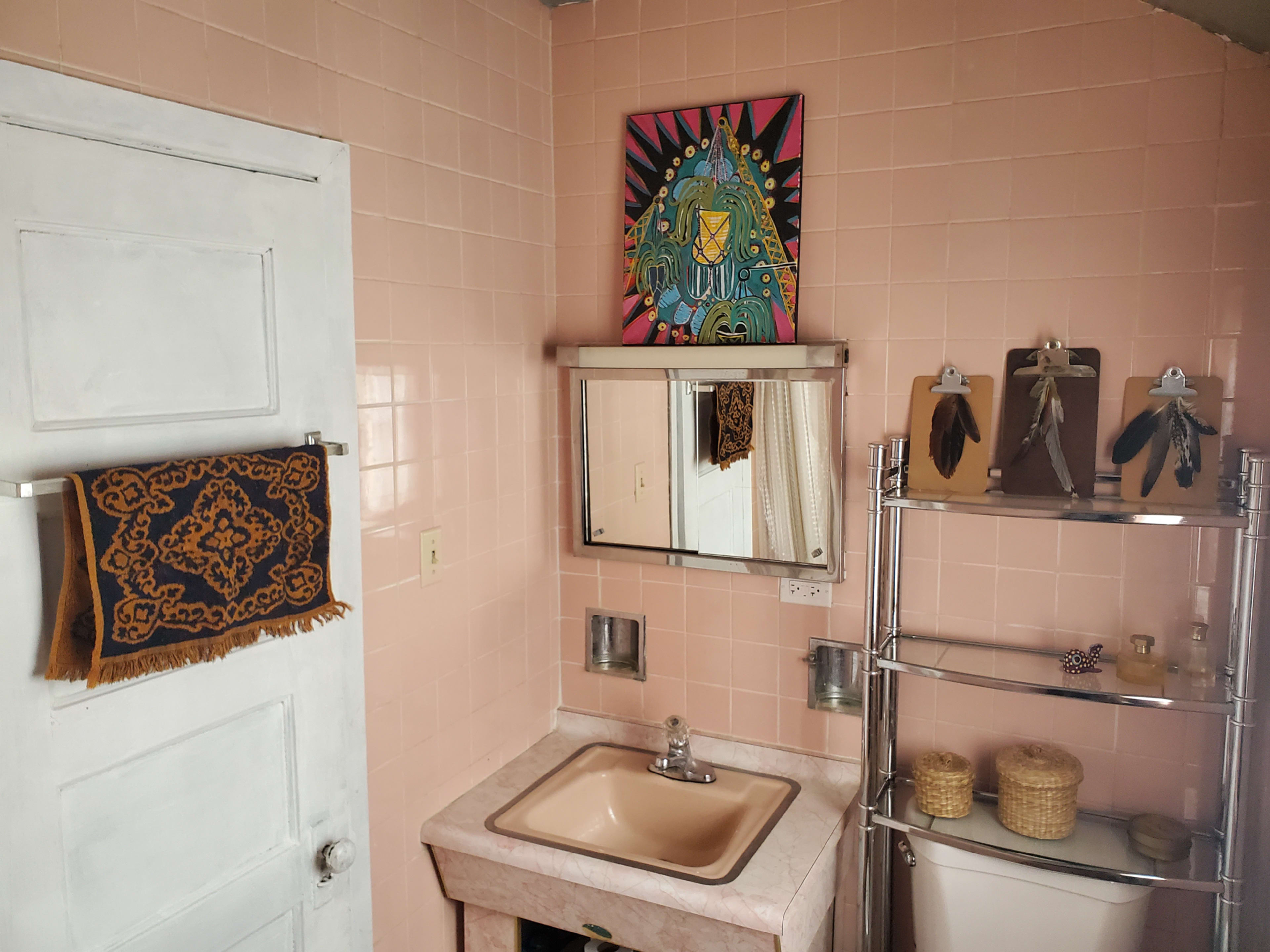 A small bathroom with pink tiled walls, a sink, a mirrored cabinet, a colorful artwork hung above, and shelves displaying decorative items.