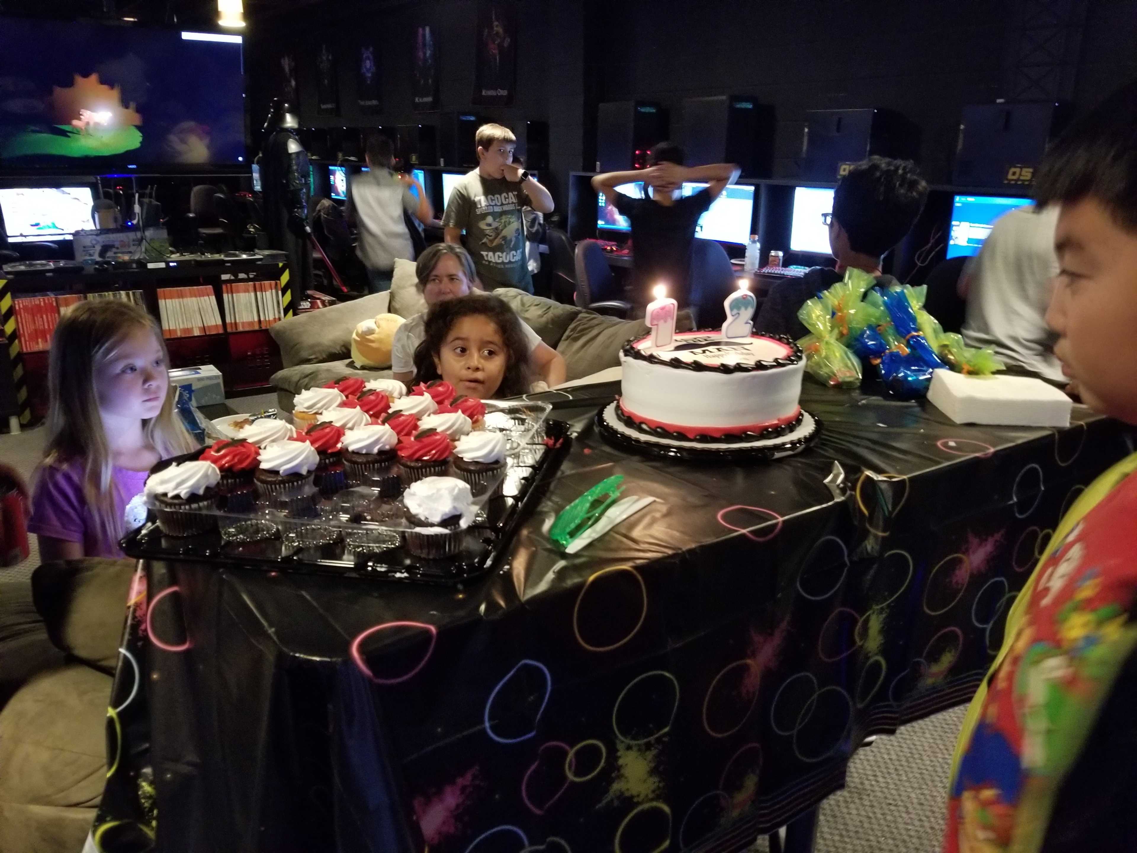 A birthday celebration takes place in a gaming lounge with a table set up featuring a decorated cake and cupcakes, surrounded by children and gaming monitors in the background.