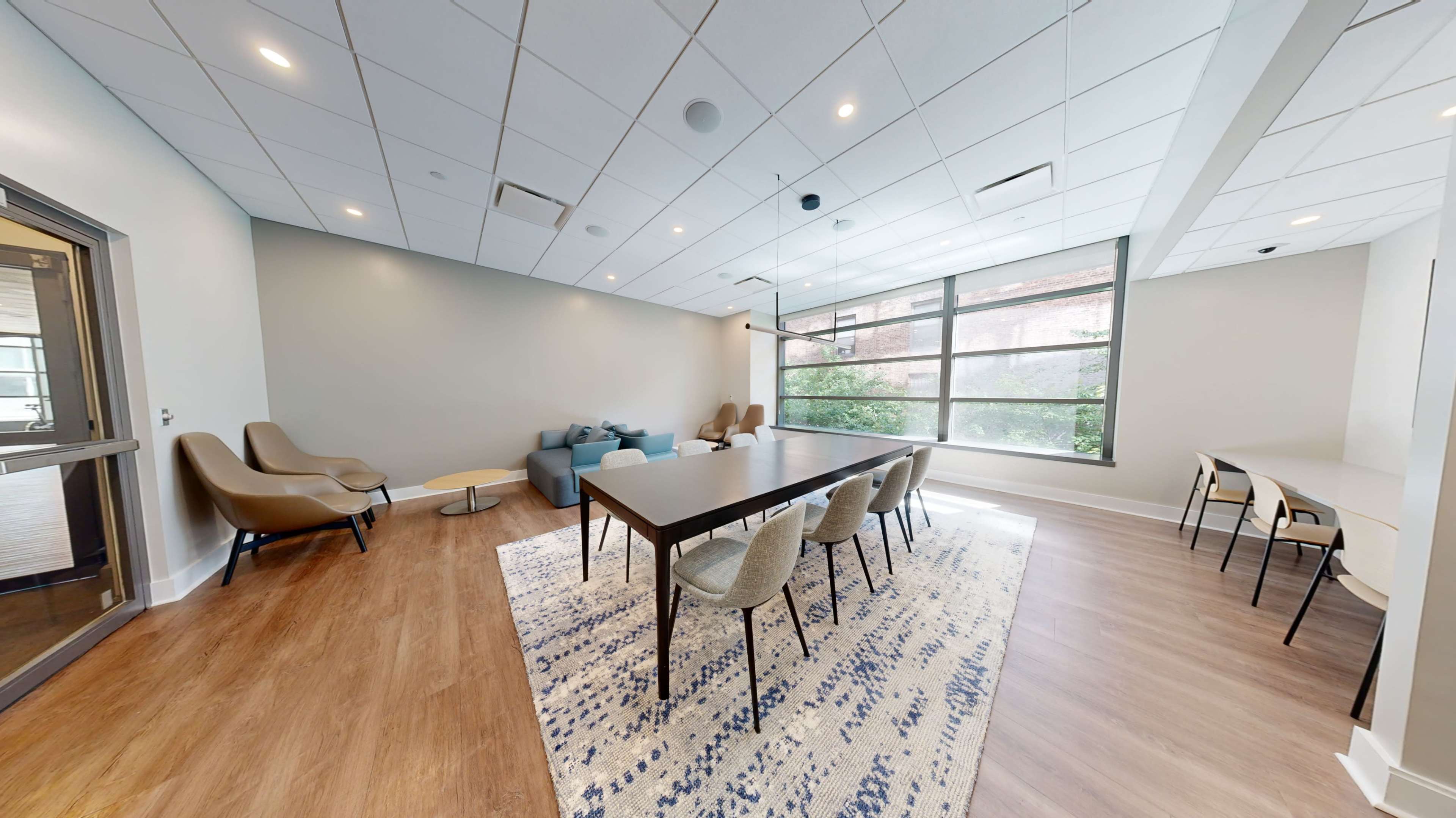 A modern lounge area features a large table, several chairs, two armchairs, a blue sofa, and large windows that allow natural light to enter the space.