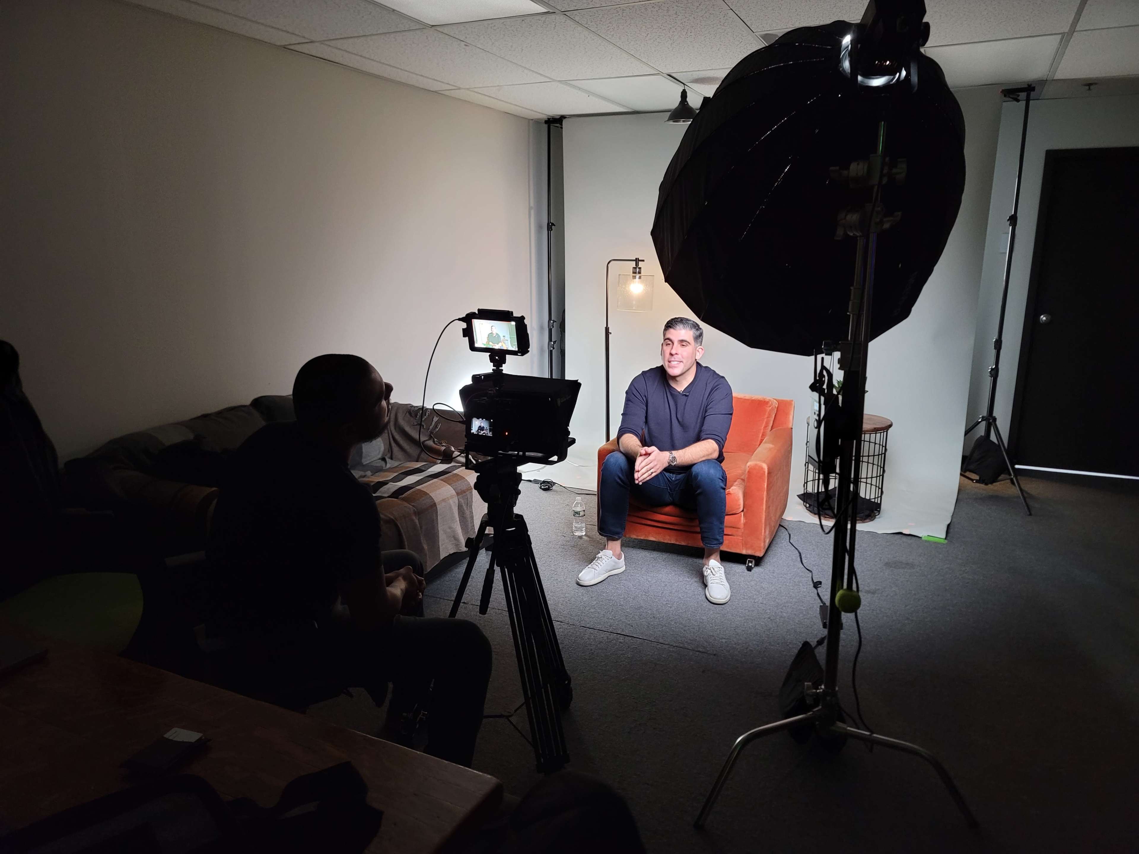 A person is being filmed while sitting on an orange couch in a dimly lit room, with a camera and lighting equipment set up in front of them.