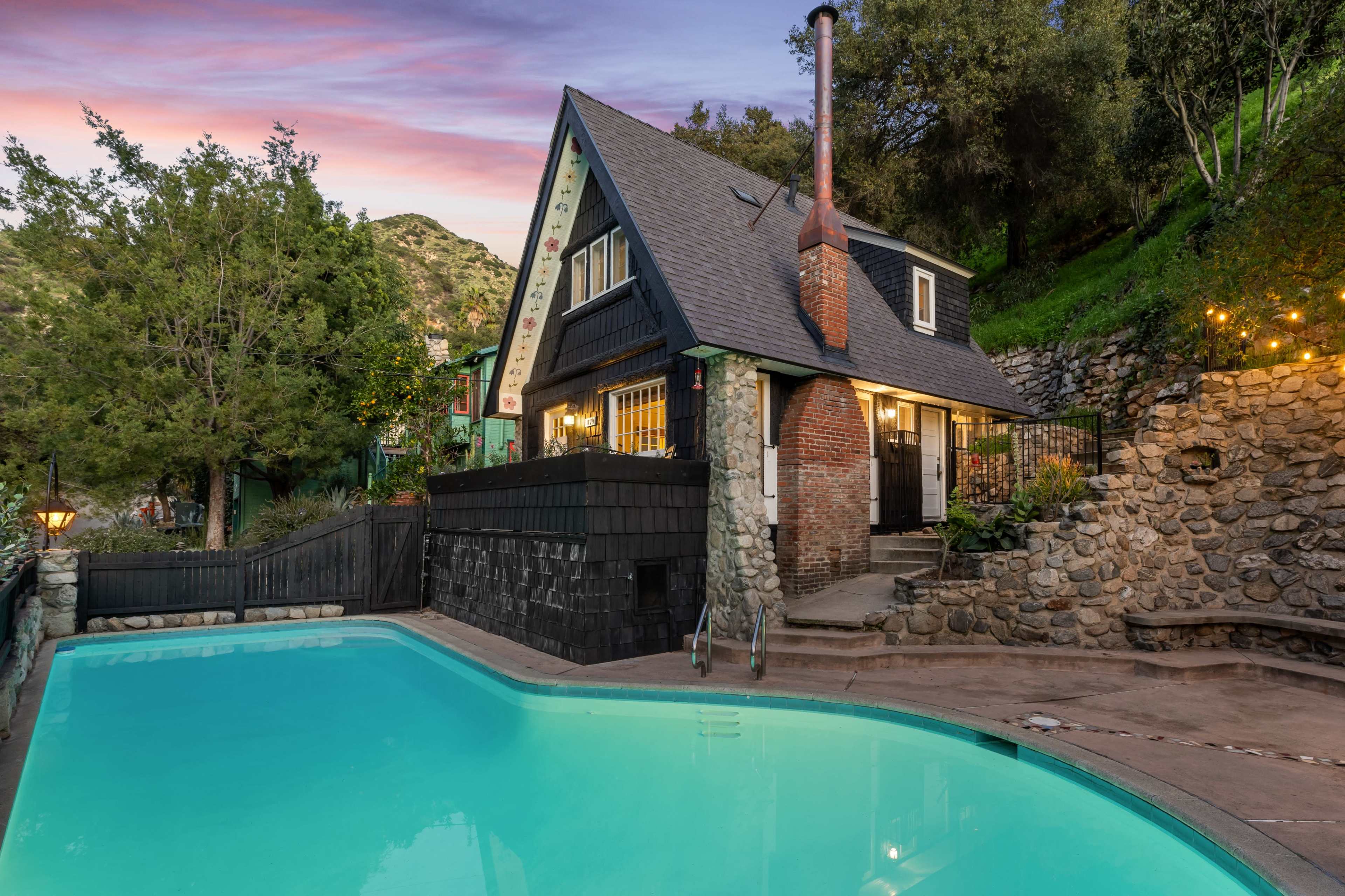 The image shows a charming house near a swimming pool, surrounded by trees and rocky terrain, with a scenic mountainous backdrop at dusk.