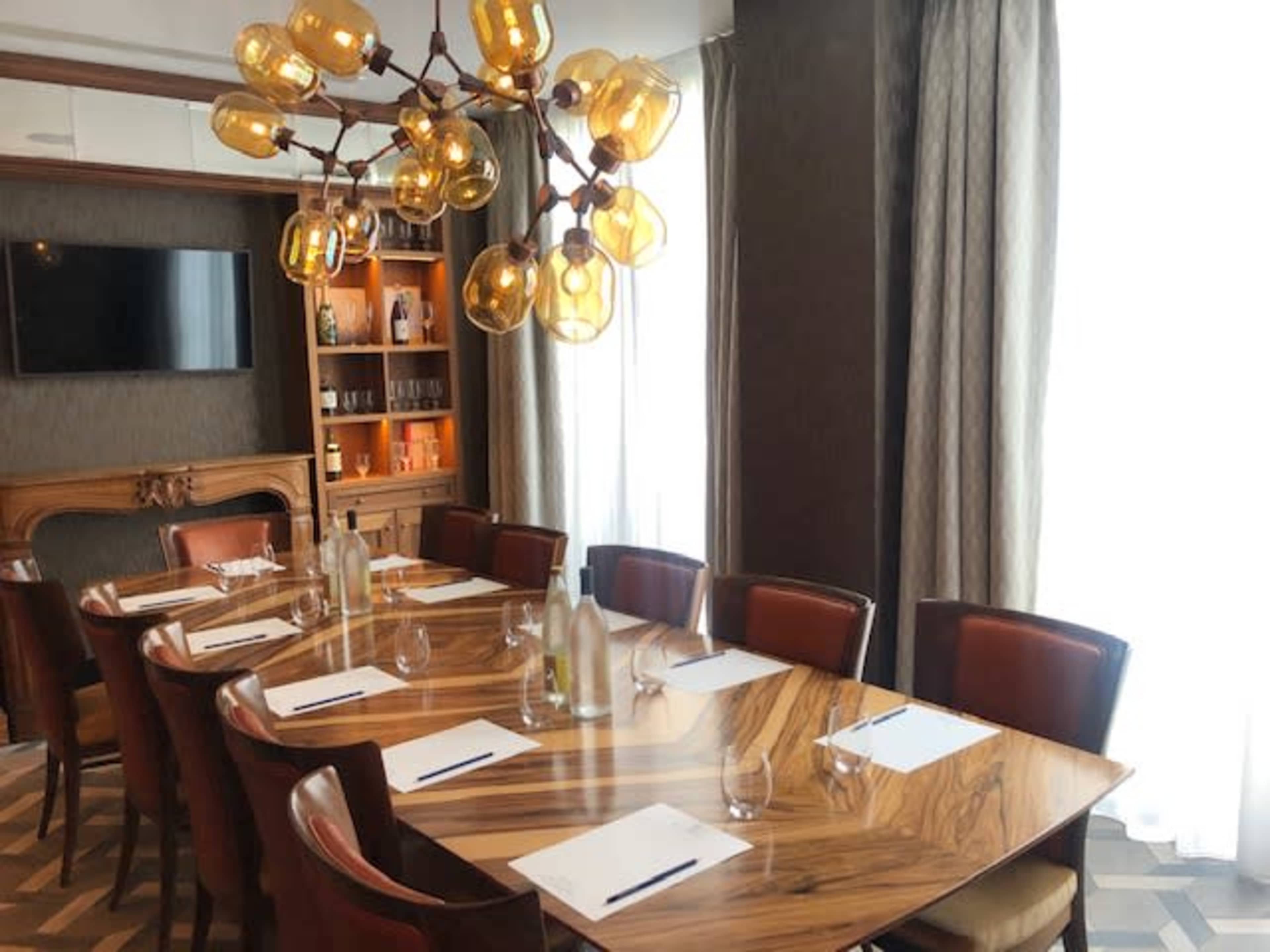 A well-lit conference room features a large wooden table set with glasses and stationery, surrounded by chairs and illuminated by a unique chandelier.