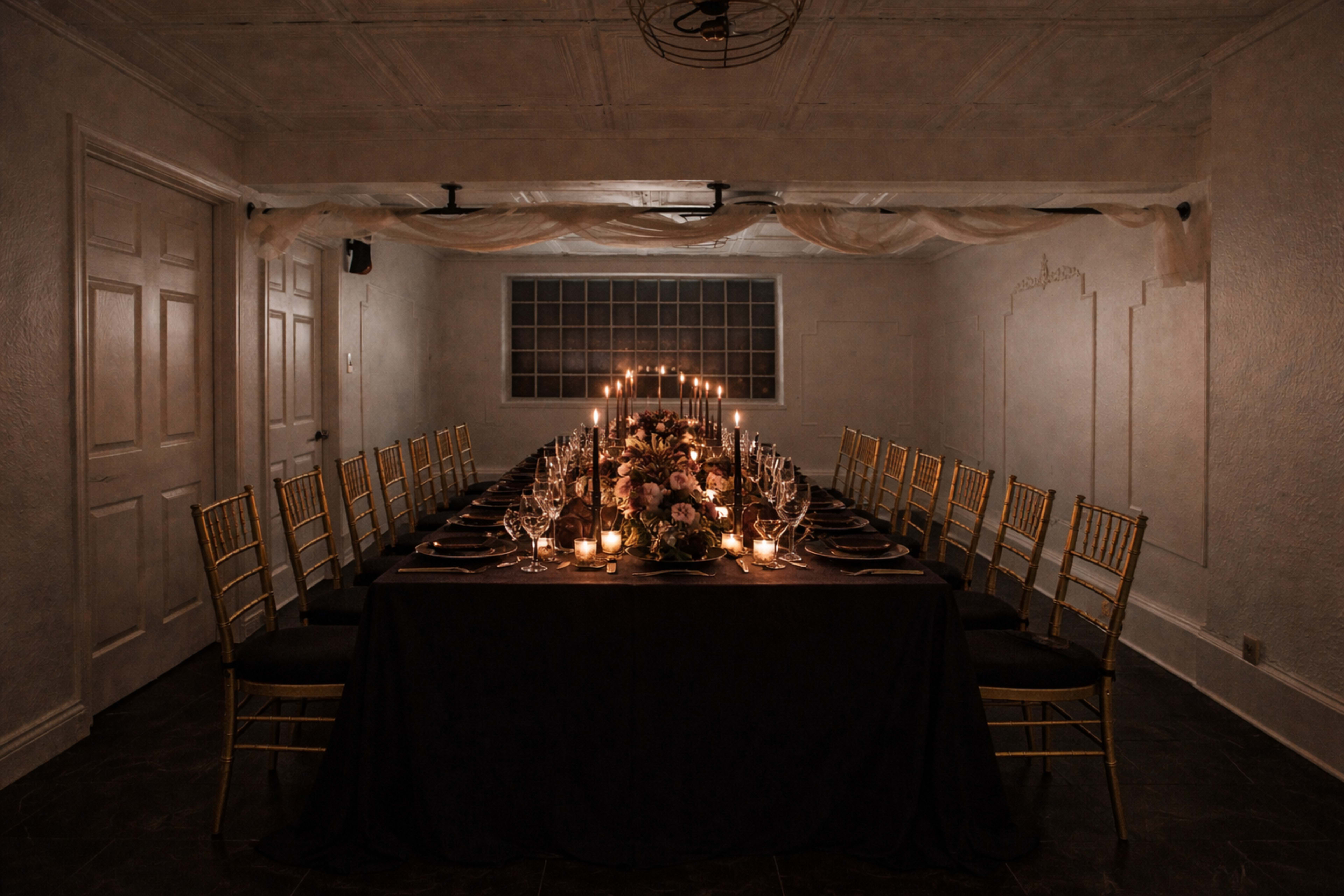 A long dining table elegantly set with candles, flowers, and tableware, is arranged in a softly lit room with white walls and a grid window.