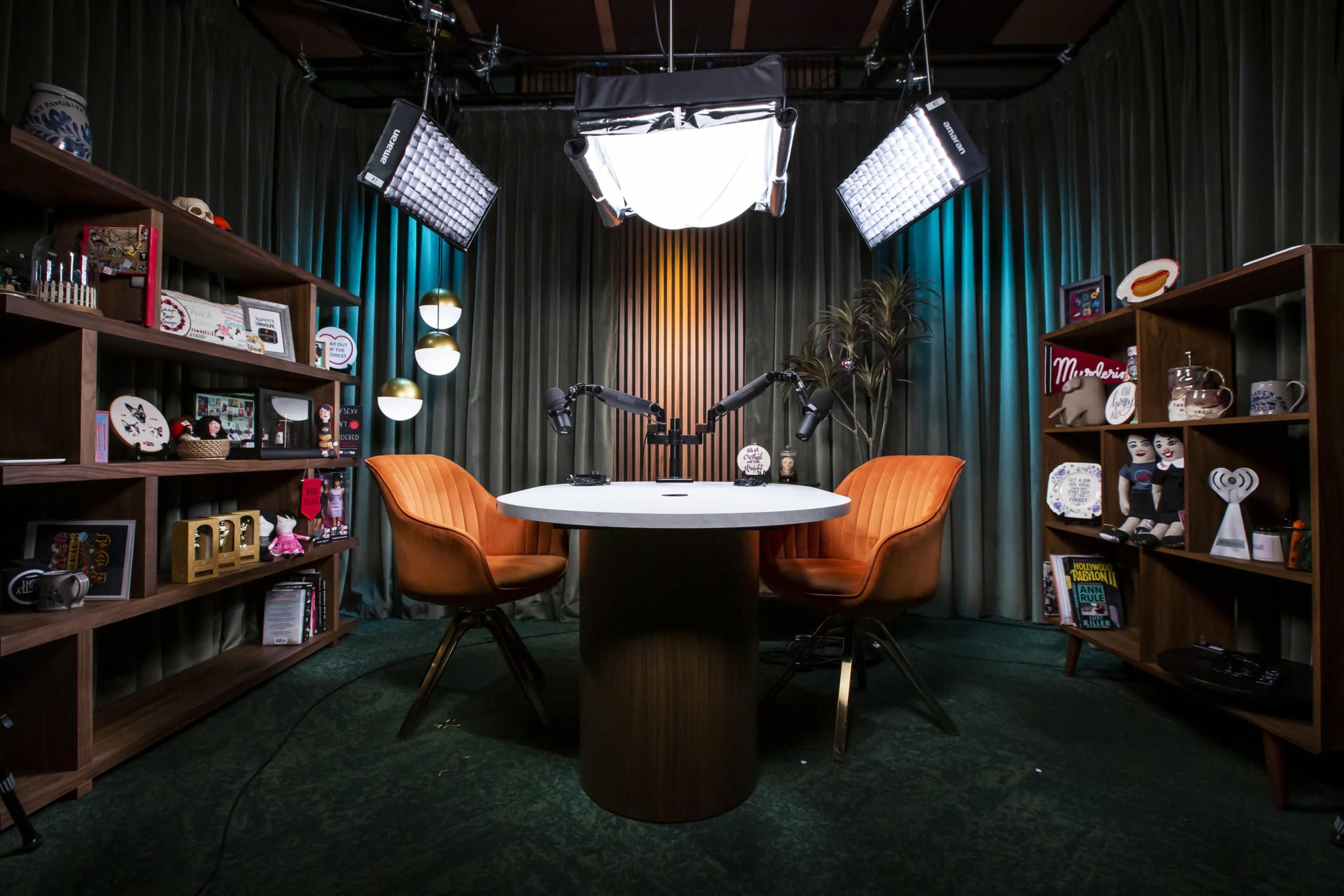 A podcast studio features two orange chairs facing a circular table, surrounded by shelves displaying various decorative items and lit by overhead lights.