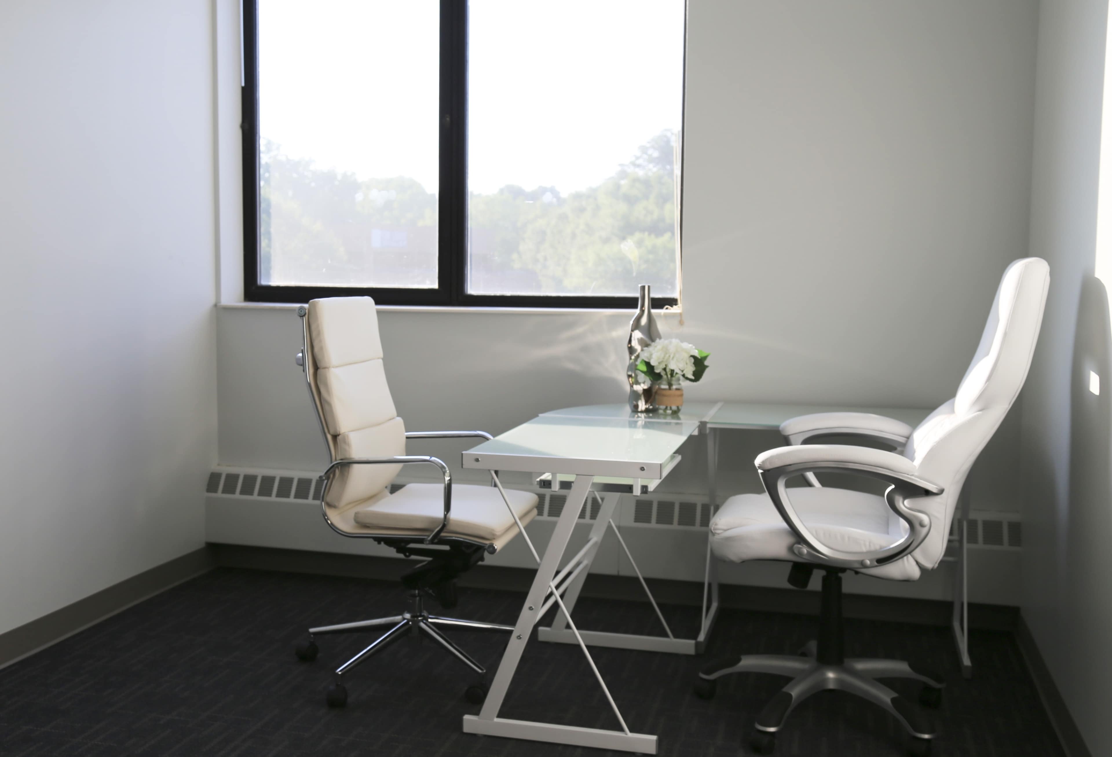 A modern office setting with two white chairs and a glass-topped desk beside a window.