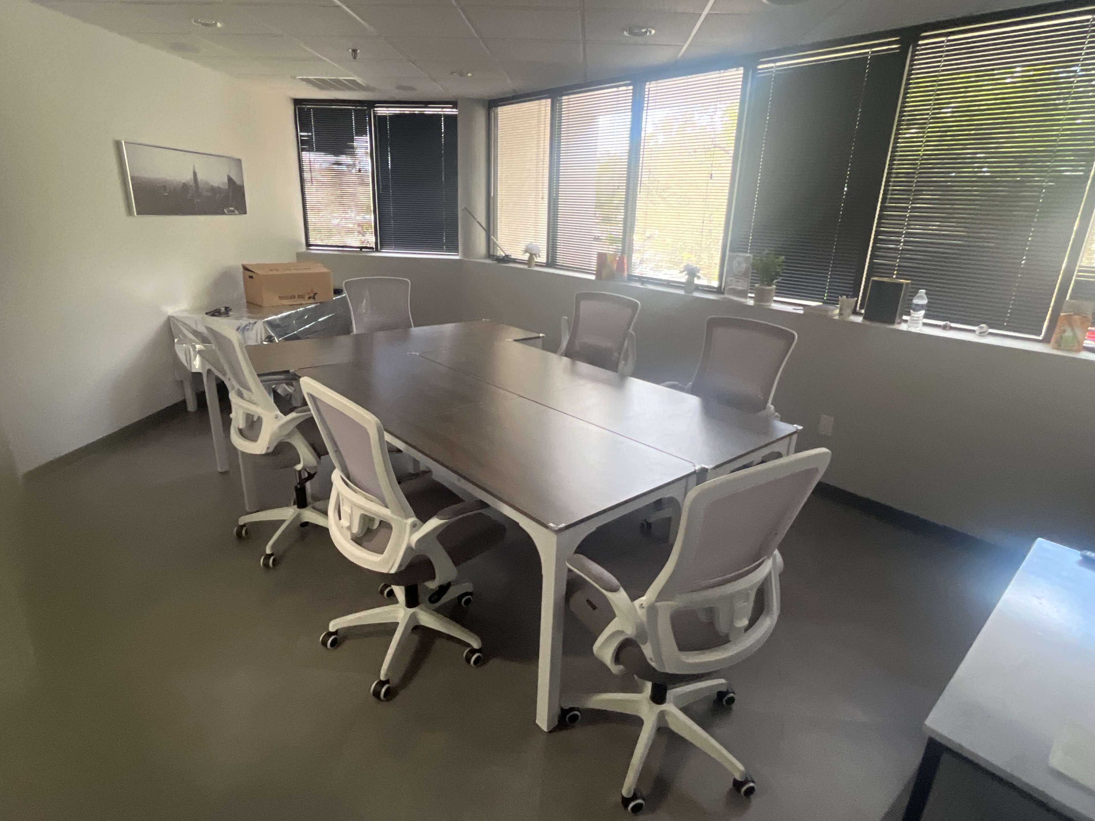 The image shows a conference room with a large table surrounded by several white office chairs, and windows letting in natural light.