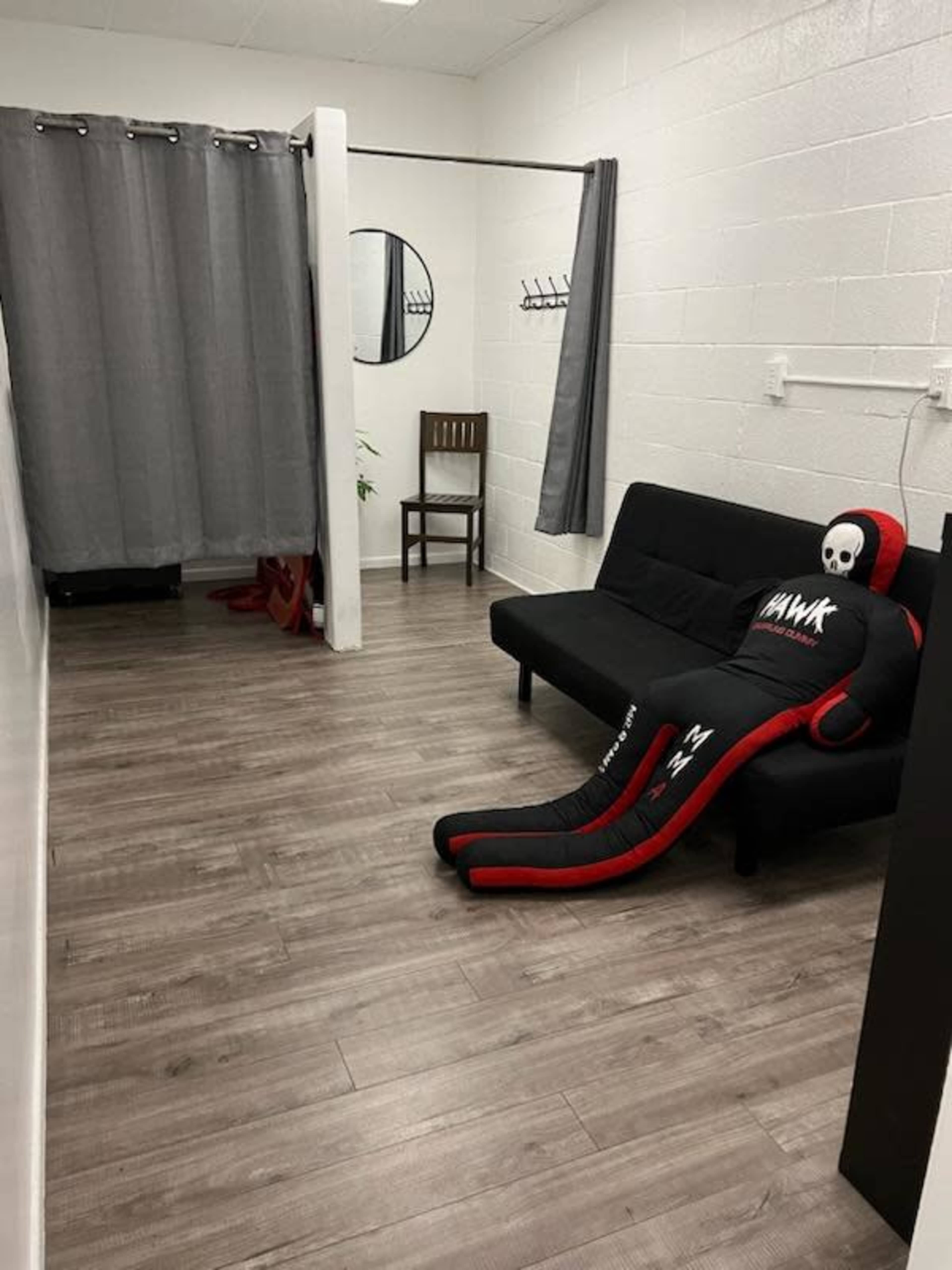A small room with a black couch and a dummy dressed in athletic gear lounging on it, alongside gray curtains and a circular mirror on the wall.