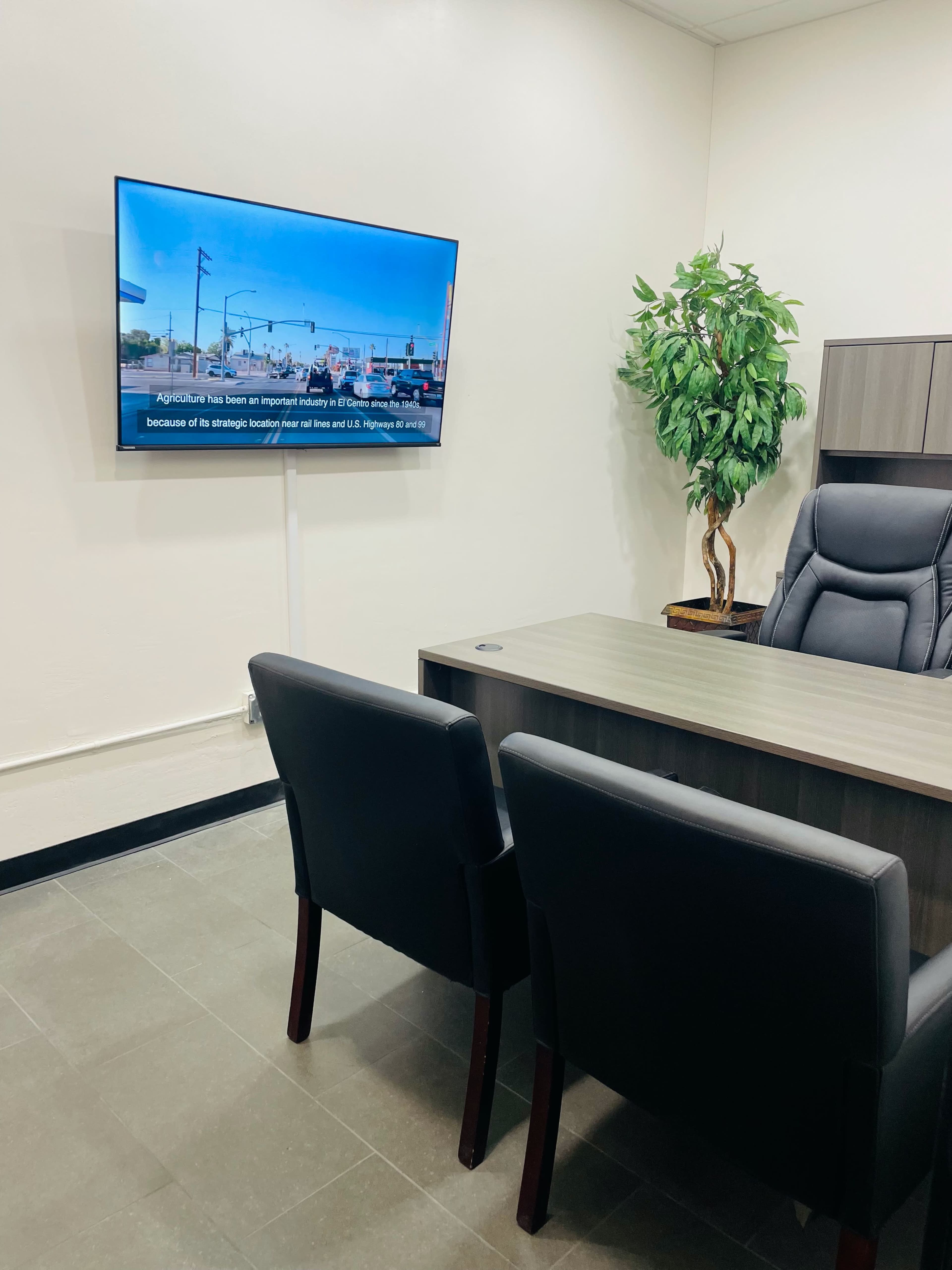 A modern office space features a wooden desk, two black chairs, and a wall-mounted television displaying a presentation.