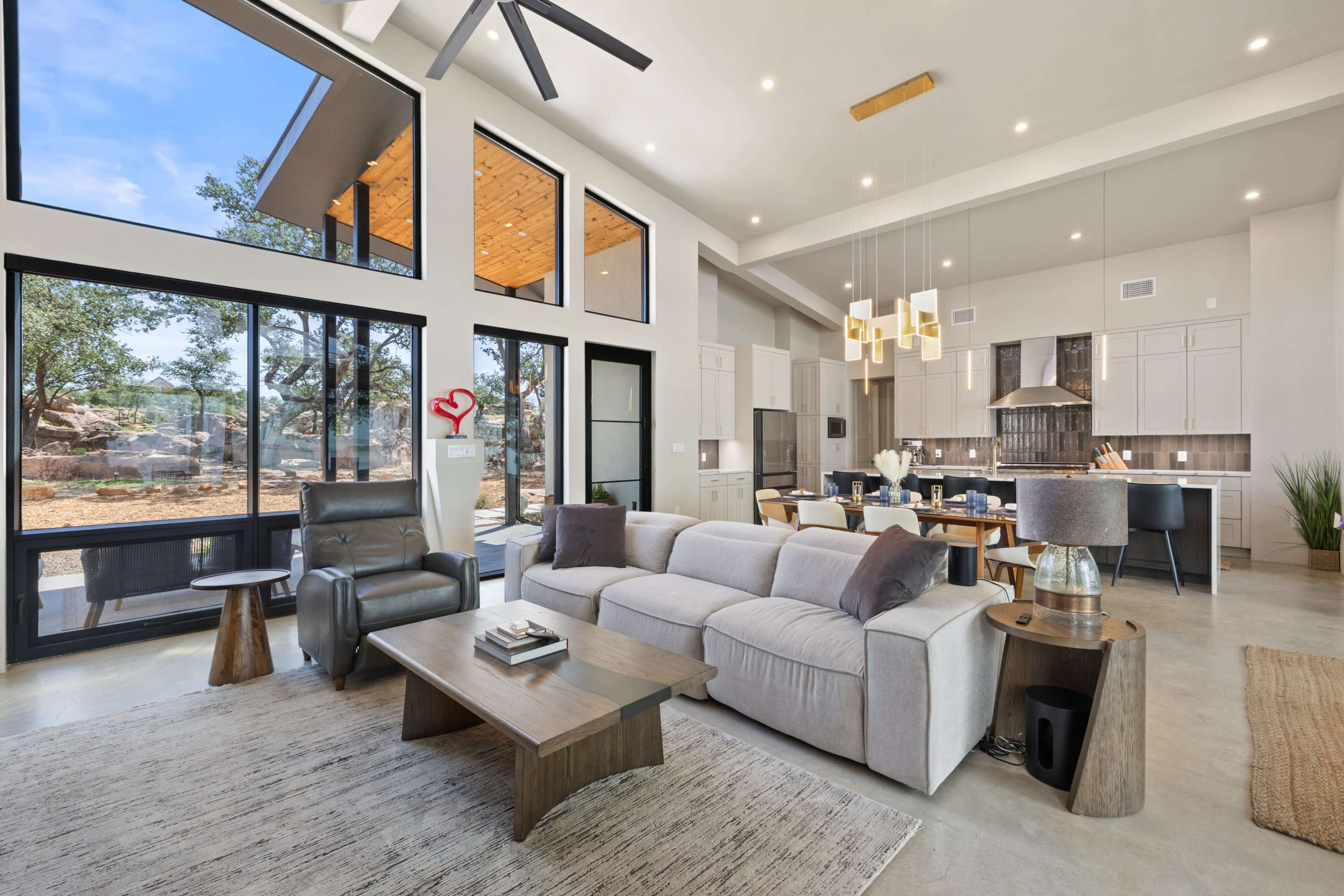 A modern living room features large windows, a sectional sofa, and a dining area that opens to a scenic outdoor space.