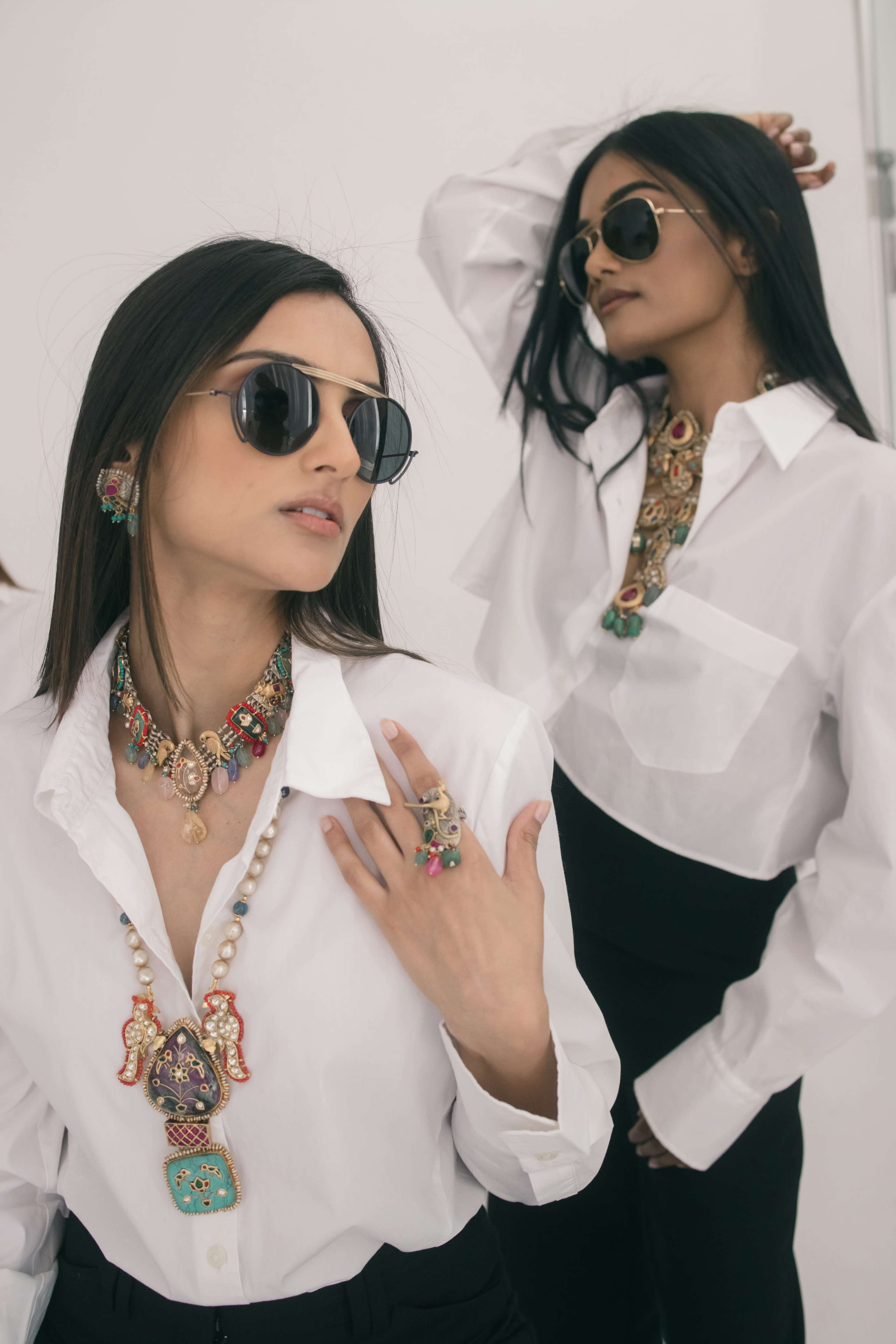 Two women wearing oversized sunglasses and white shirts, adorned with colorful jewelry, pose against a minimalistic background.
