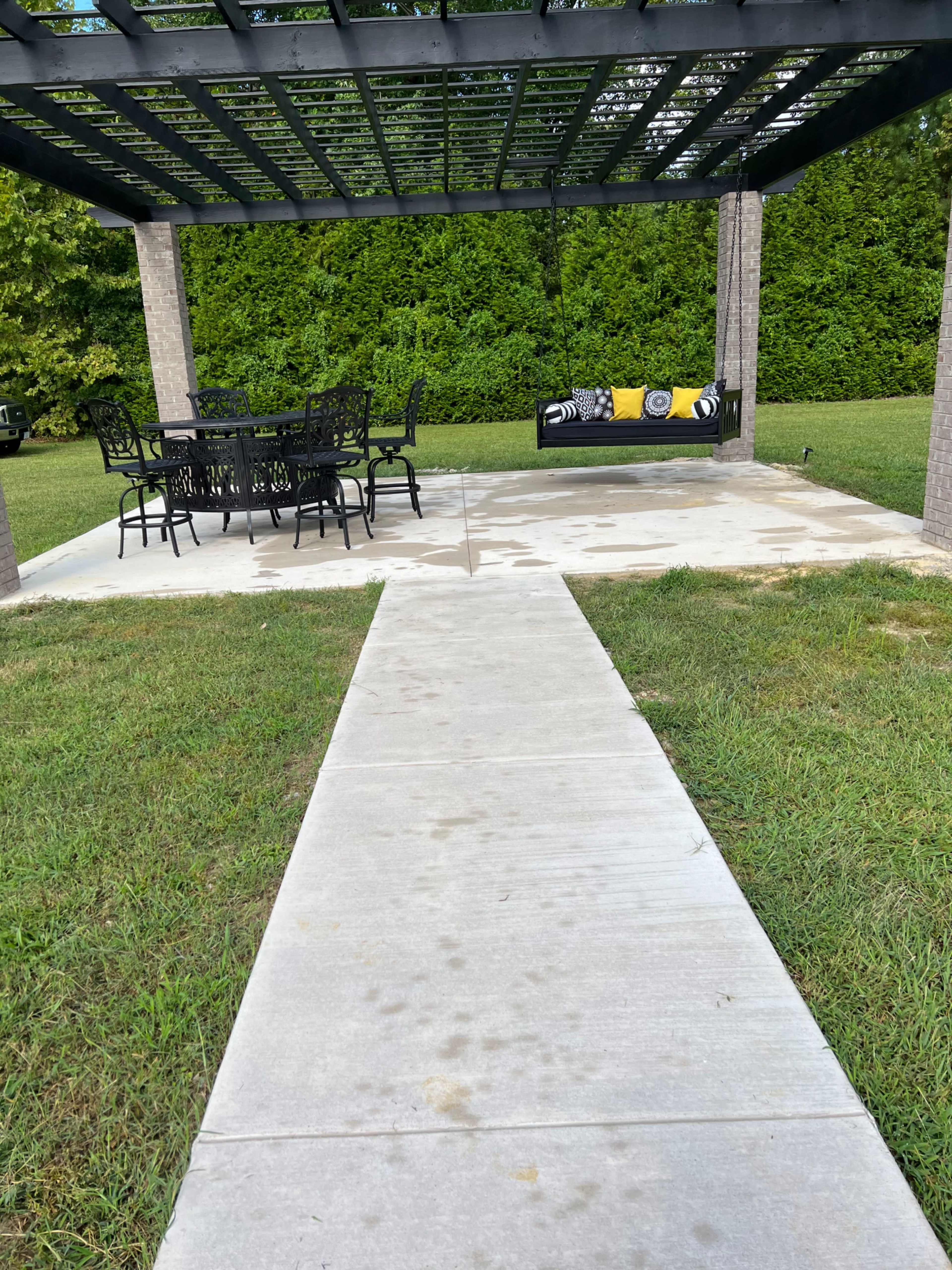 A concrete pathway leads to a covered patio featuring a dining table and a hanging bench surrounded by grass.