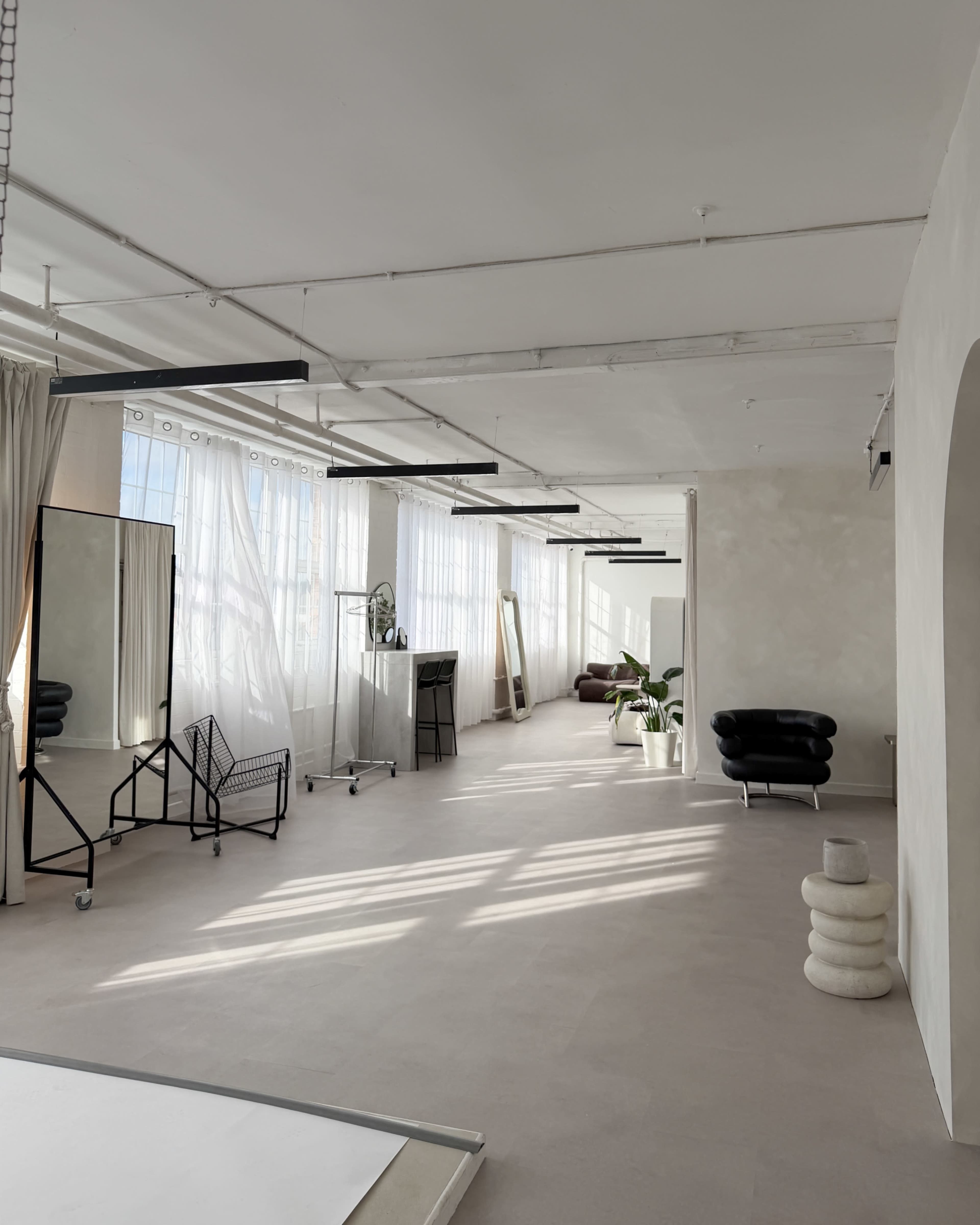 The image shows a bright, airy studio space with large windows, light-colored walls, and minimalist furniture arrangements including a black chair and a mirror stand.