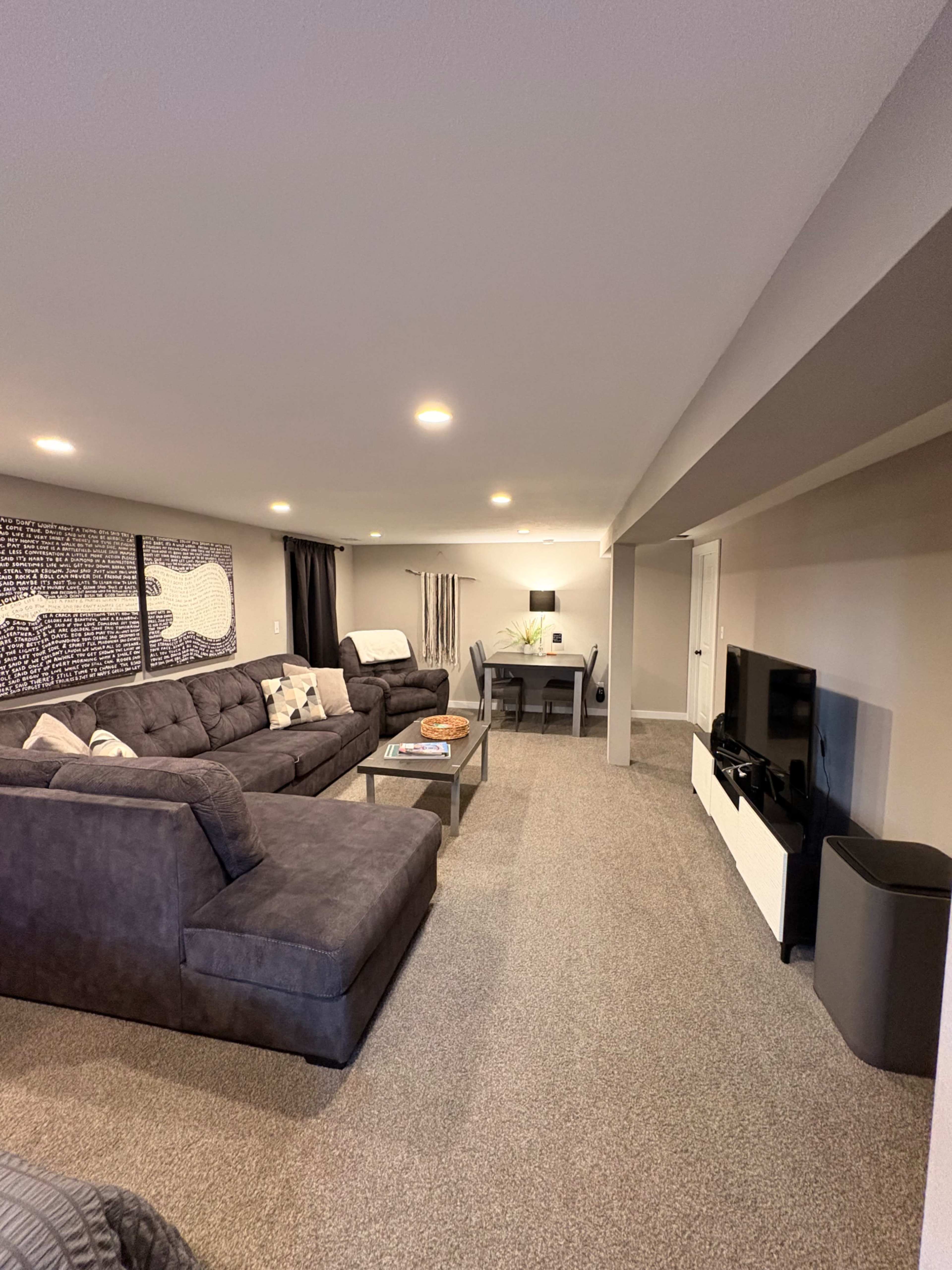 The image shows a finished basement living area featuring a large sectional sofa, a coffee table, a dining table, and a wall-mounted television.