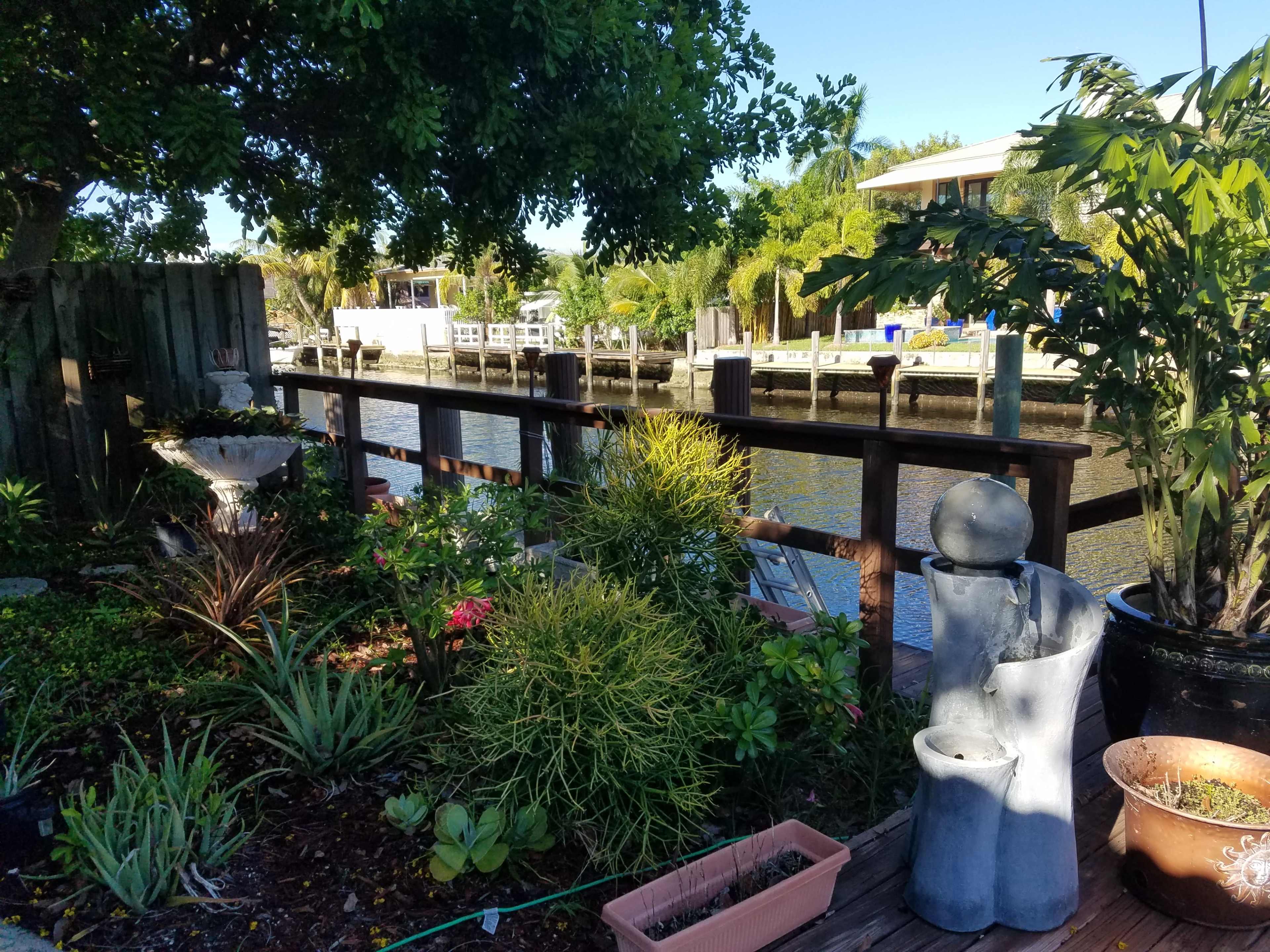 The image shows a canal with a wooden dock alongside a garden featuring various plants and a stone statue.