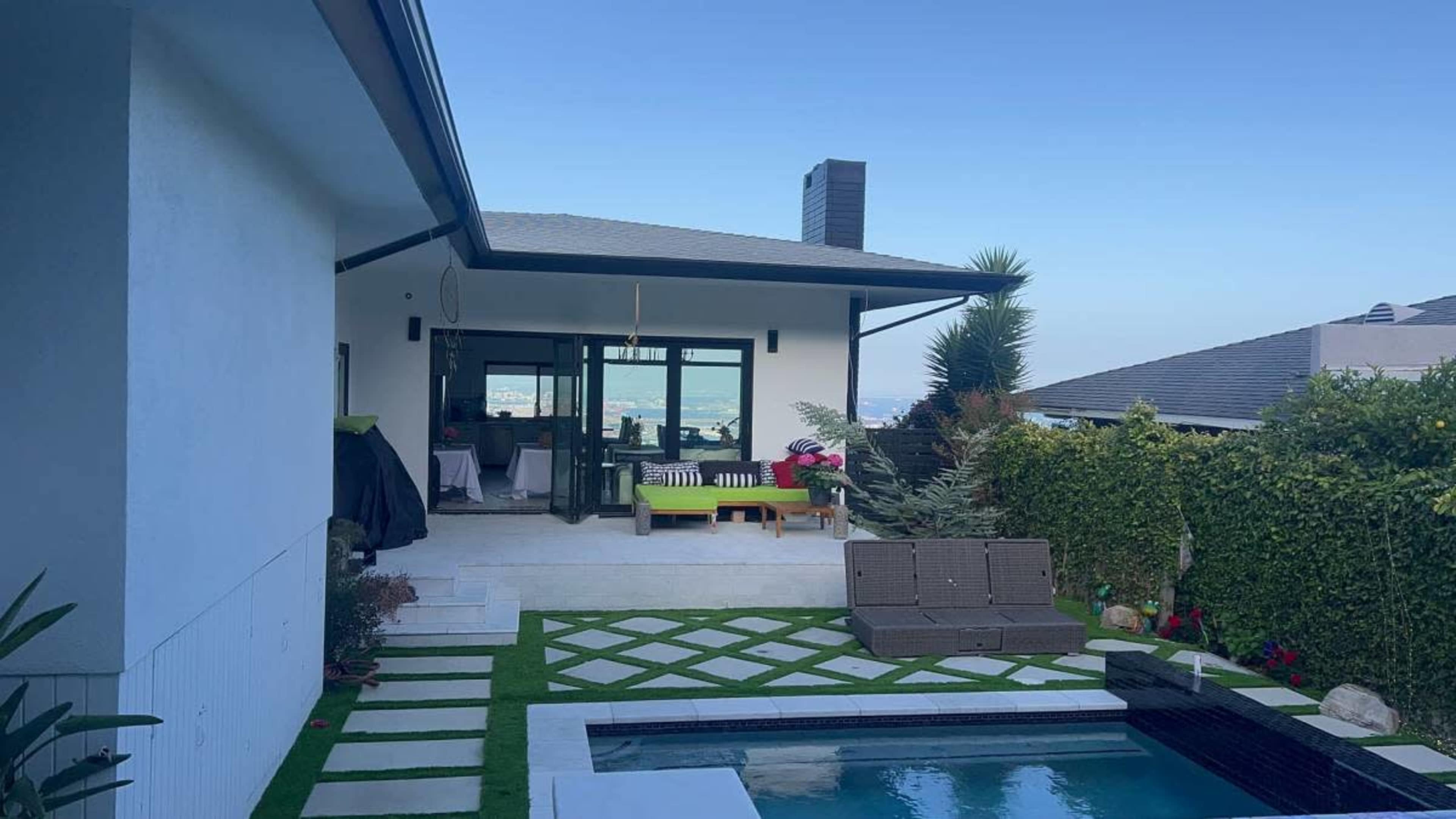 The image features a modern backyard with a swimming pool, a patio area with outdoor furniture, and a view of a nearby body of water in the background.