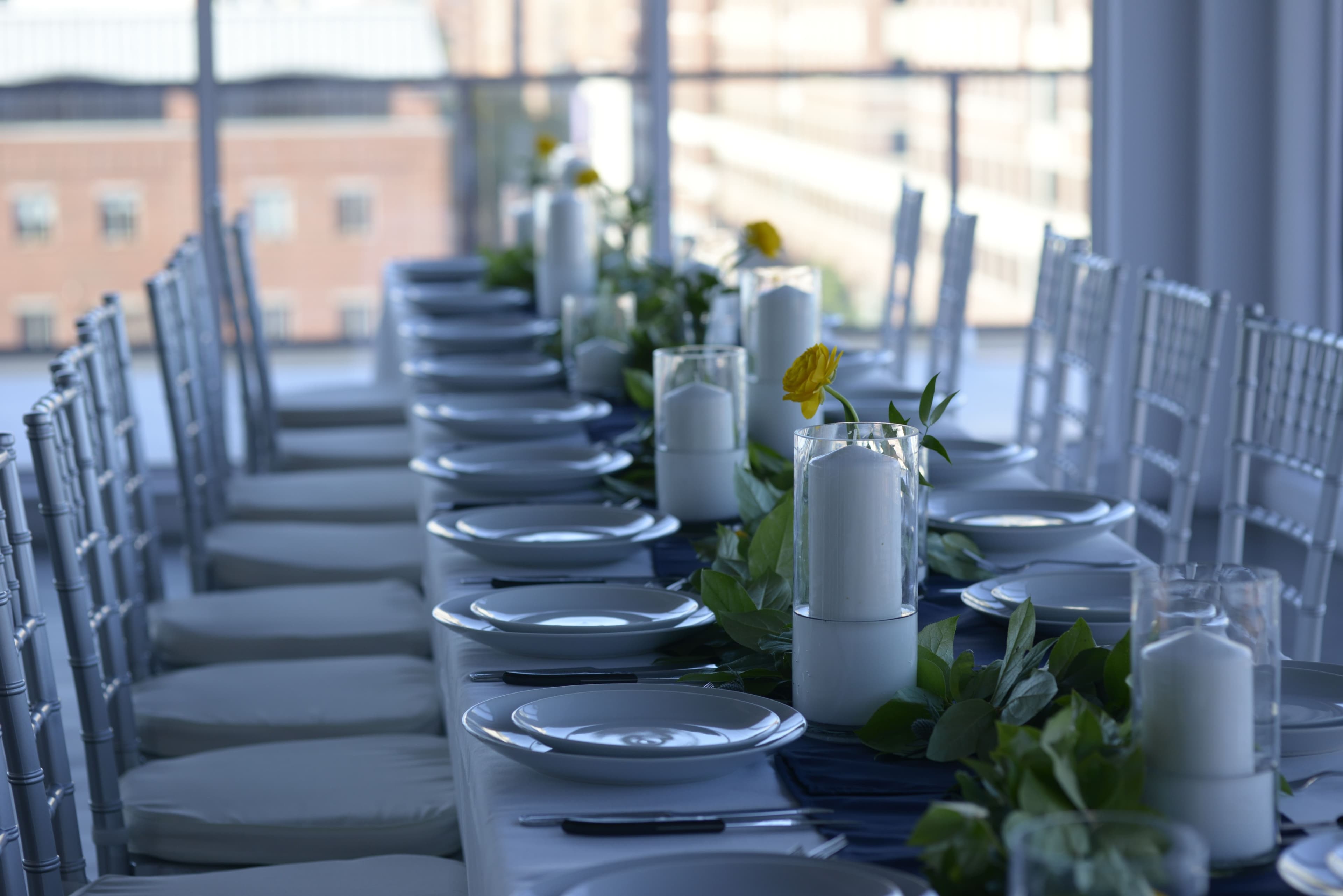 A long banquet table is set with white plates, glass vases containing candles, and greenery, arranged in a bright room with large windows overlooking a cityscape.
