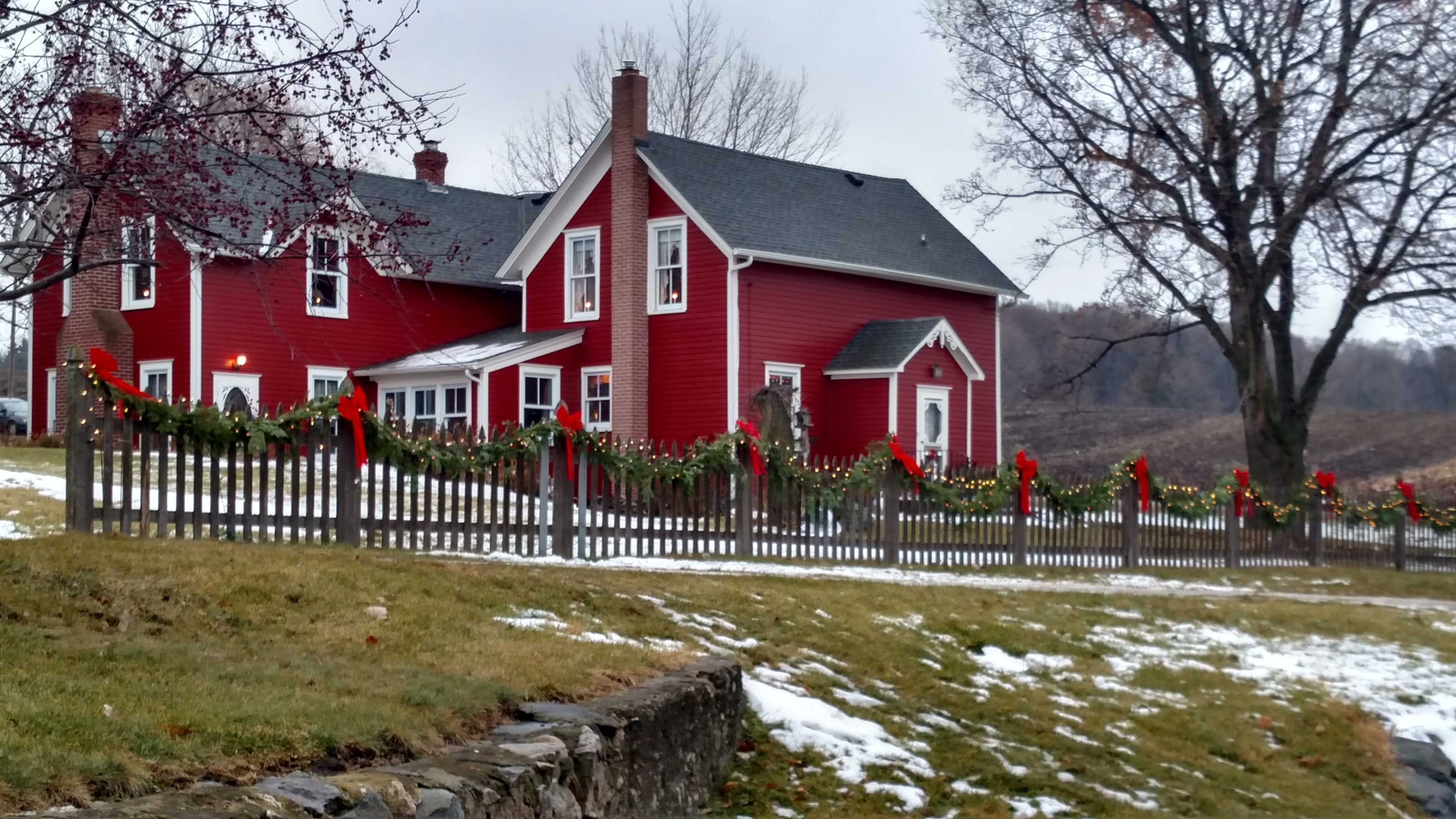 A red house with white trim is decorated with garlands and bows along a picket fence, surrounded by a snowy yard and bare trees.