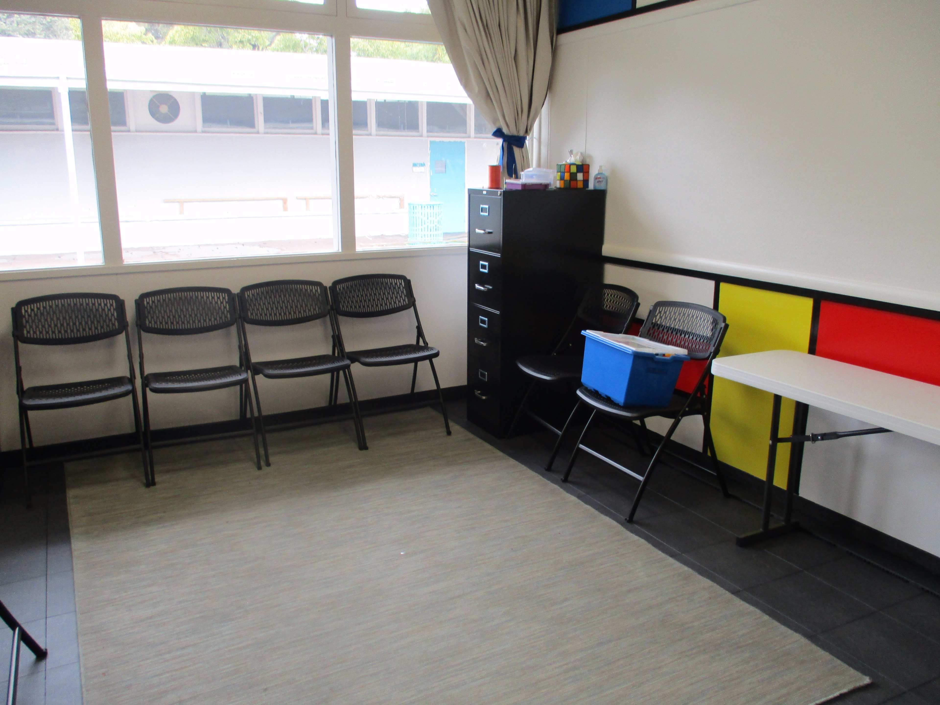 A classroom with a few black chairs arranged in a semi-circle, a filing cabinet, a blue container on top of it, and a white table against a colorful wall.