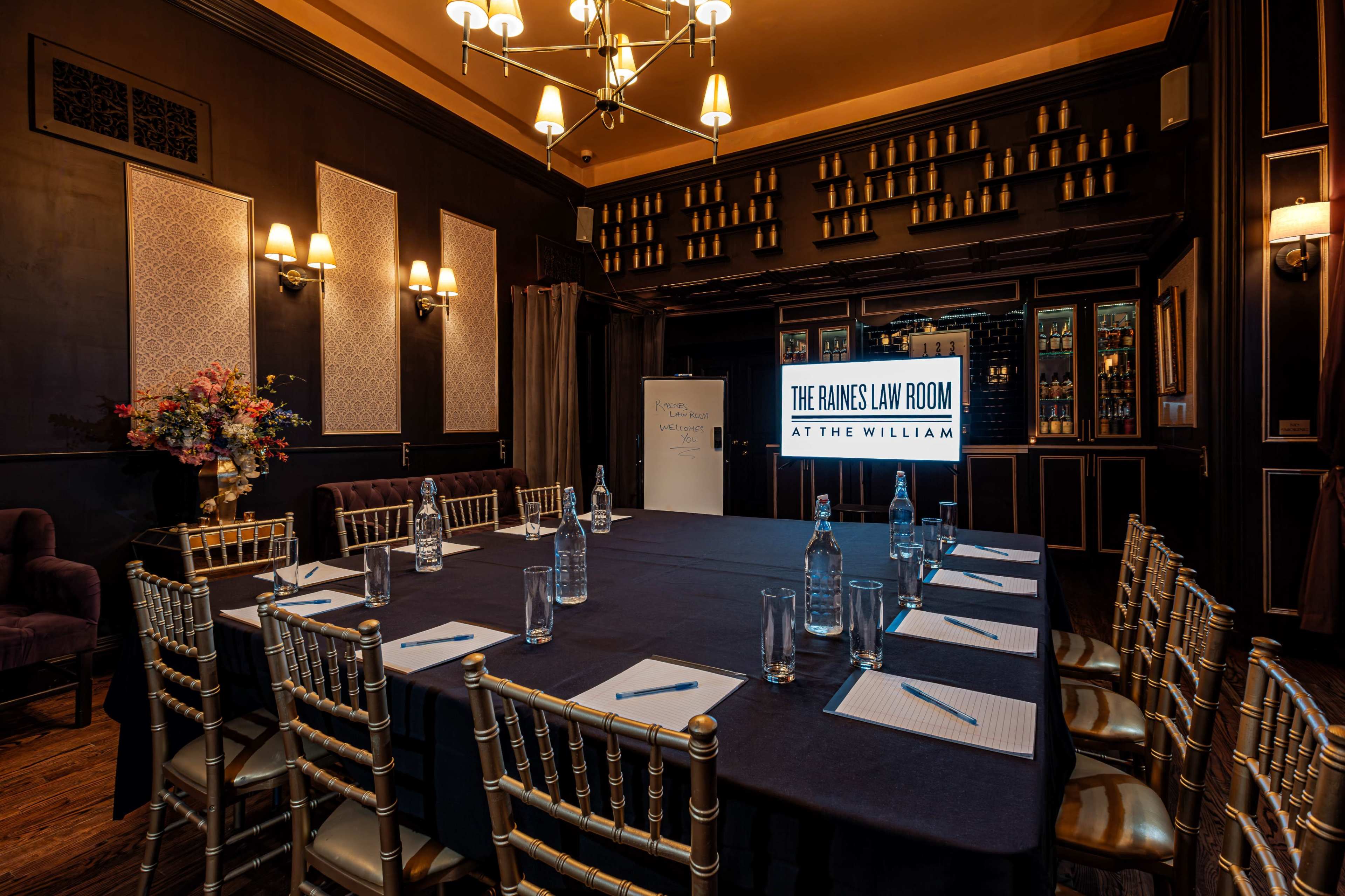The image shows a conference room set up with a large table, chairs, and a screen displaying "The Raines Law Room at The William," surrounded by shelves of bottles in a dark, elegant interior.