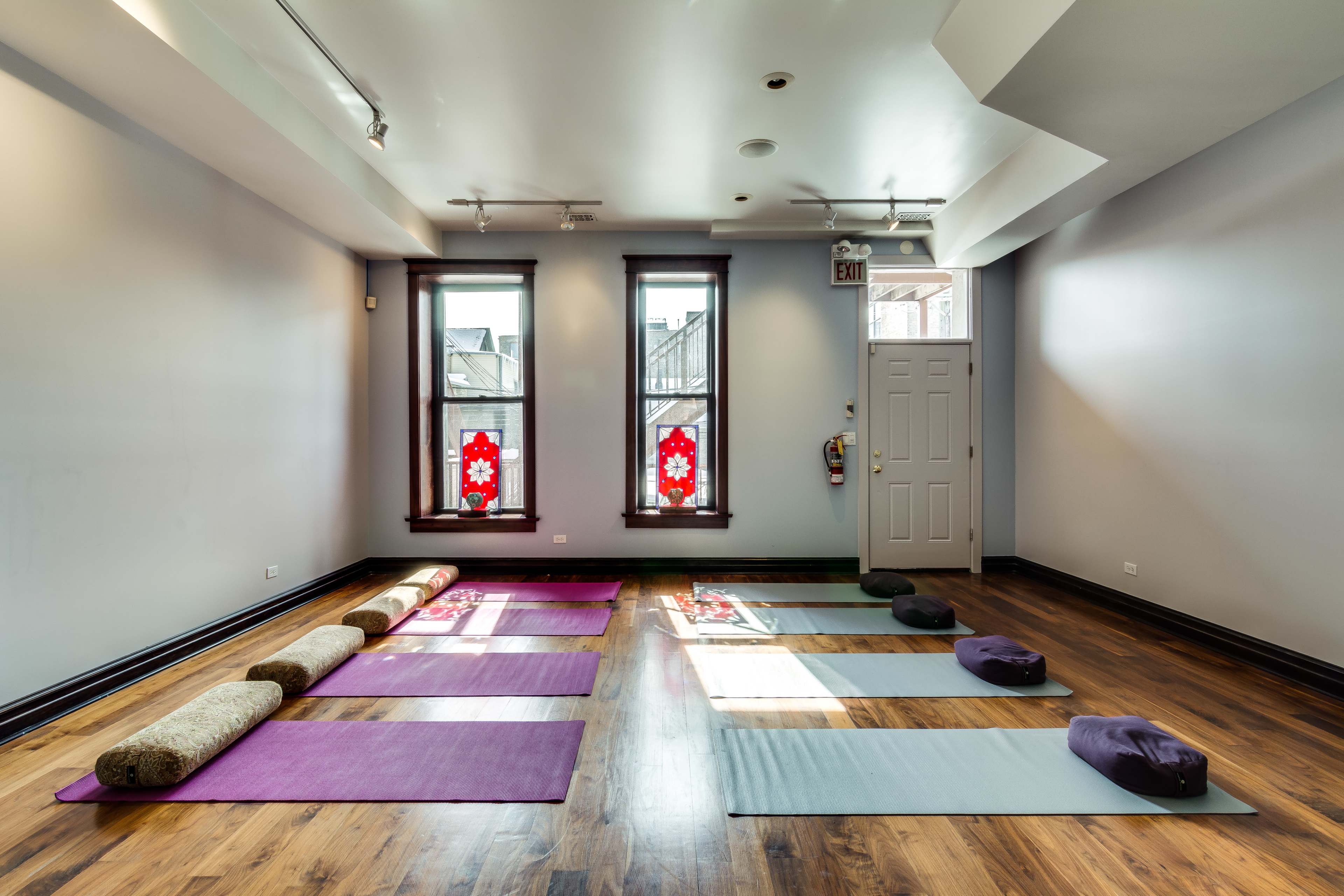 A yoga studio with several yoga mats arranged on a wooden floor, and two windows featuring red decorative elements.