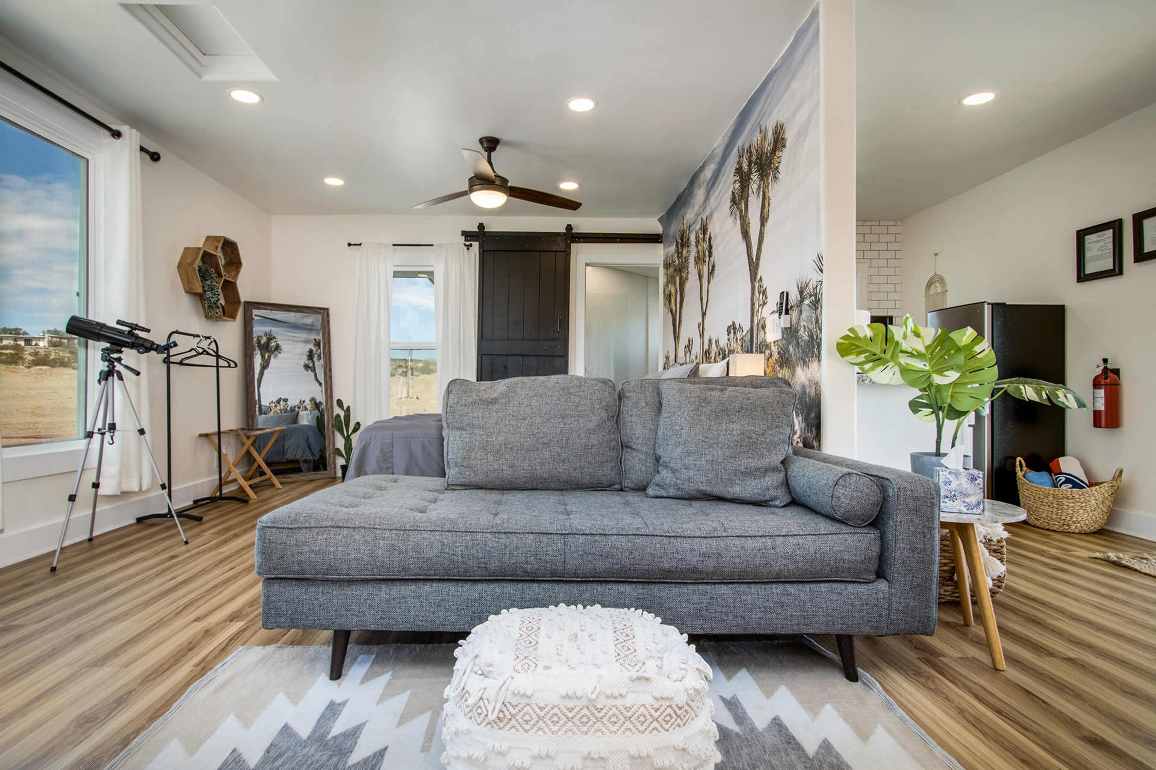 The image shows a modern living room featuring a gray couch, a decorative wall with a landscape mural, and various furnishings including a tripod and a potted plant.