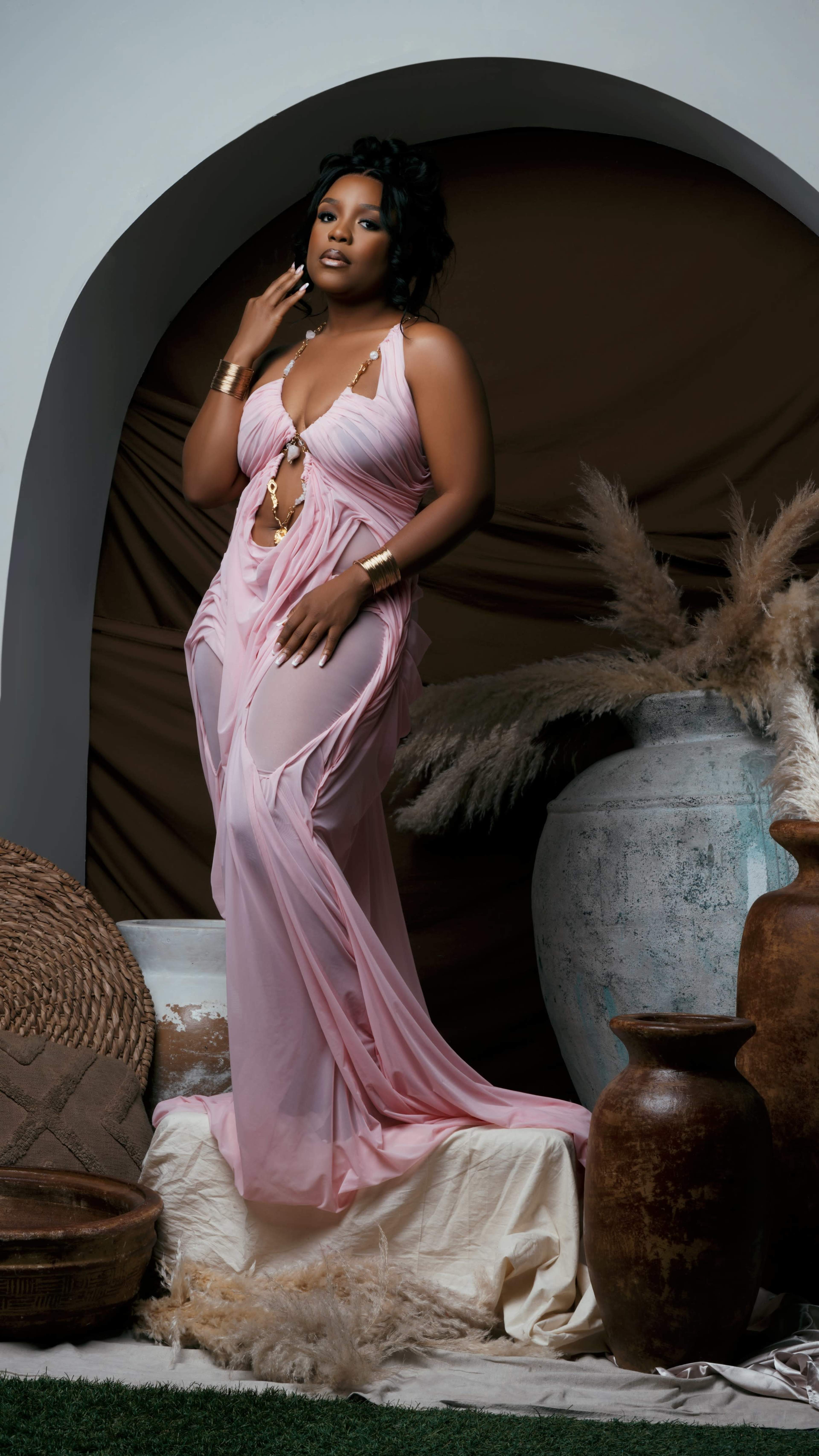 A woman in a pink gown poses elegantly among decorative pottery and woven baskets.