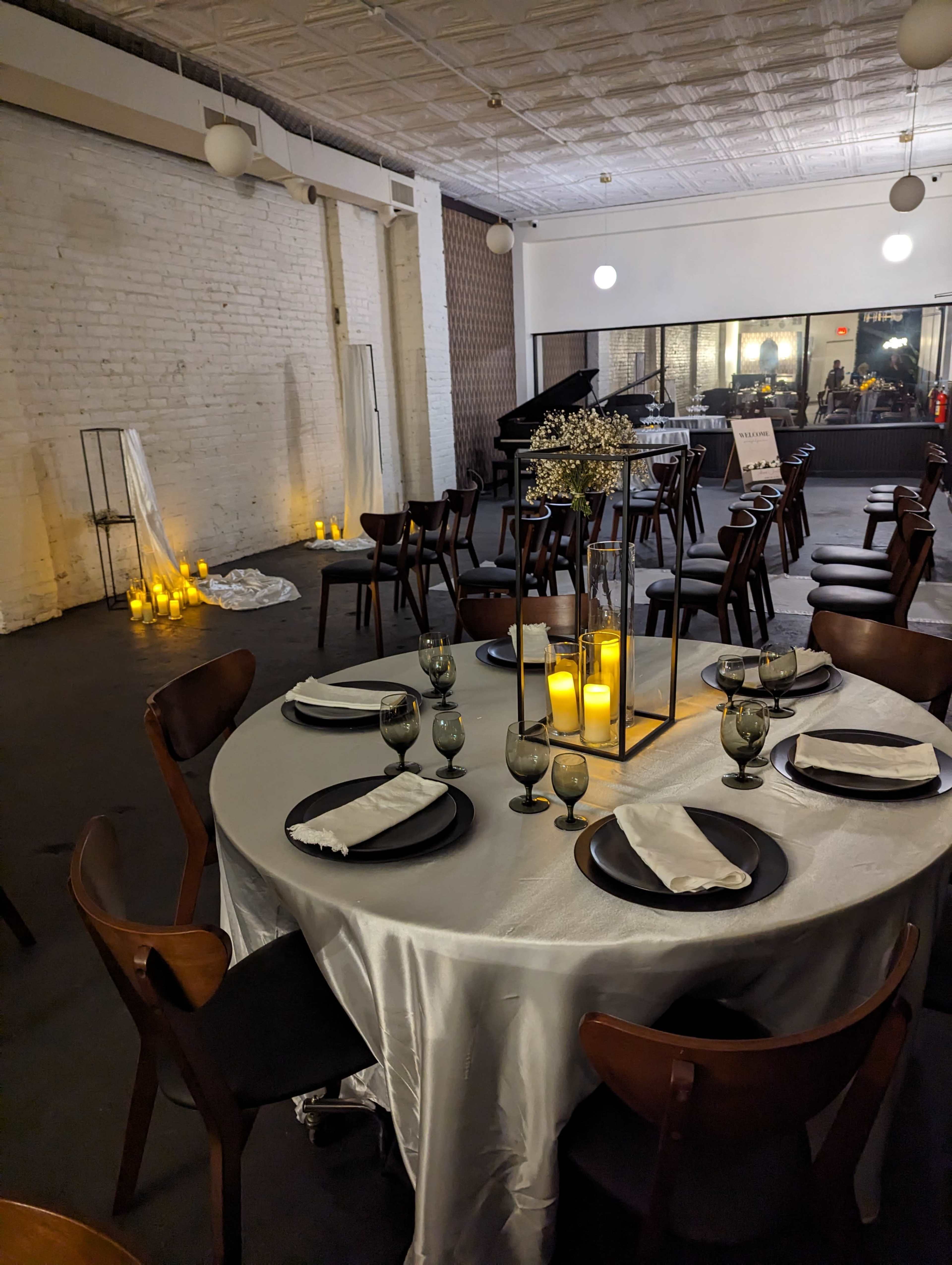 A dining area is set with a round table featuring a gray tablecloth, glassware, and candle centerpieces, while soft lighting and candles illuminate the background.