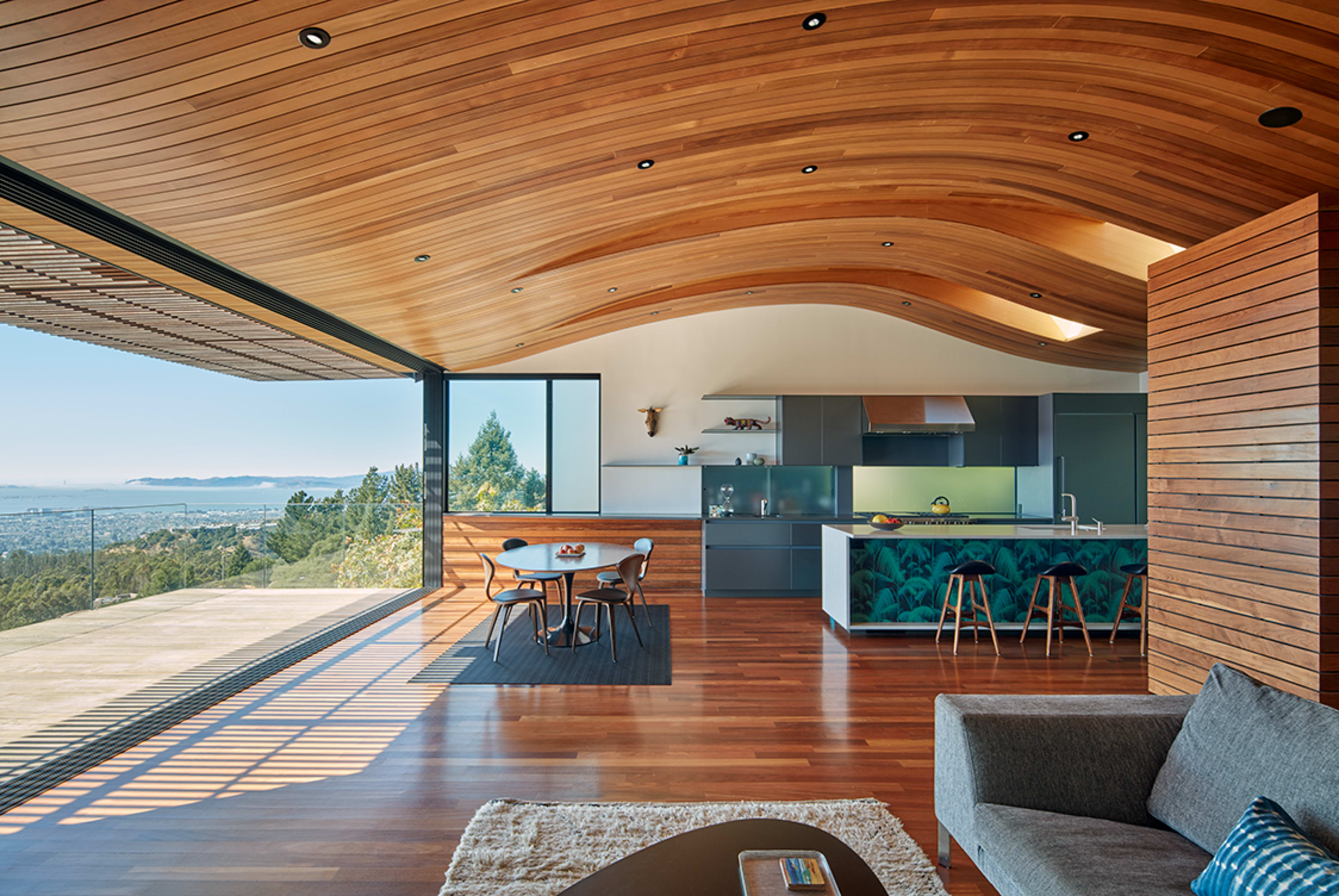 A modern interior features a spacious living area with a kitchen, large windows offering a view, and a wooden ceiling.