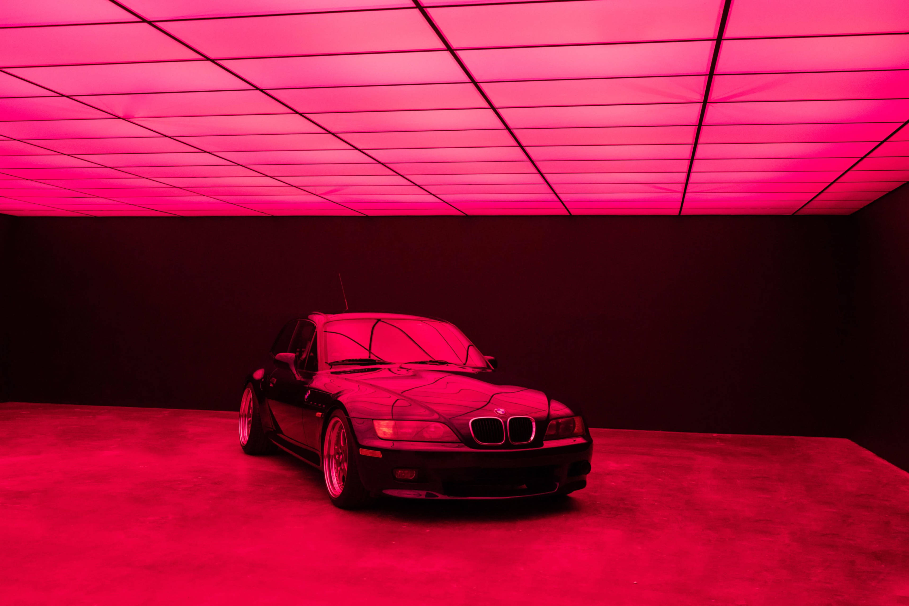 A black BMW sits under bright pink lighting in a minimalist room with a flat ceiling.