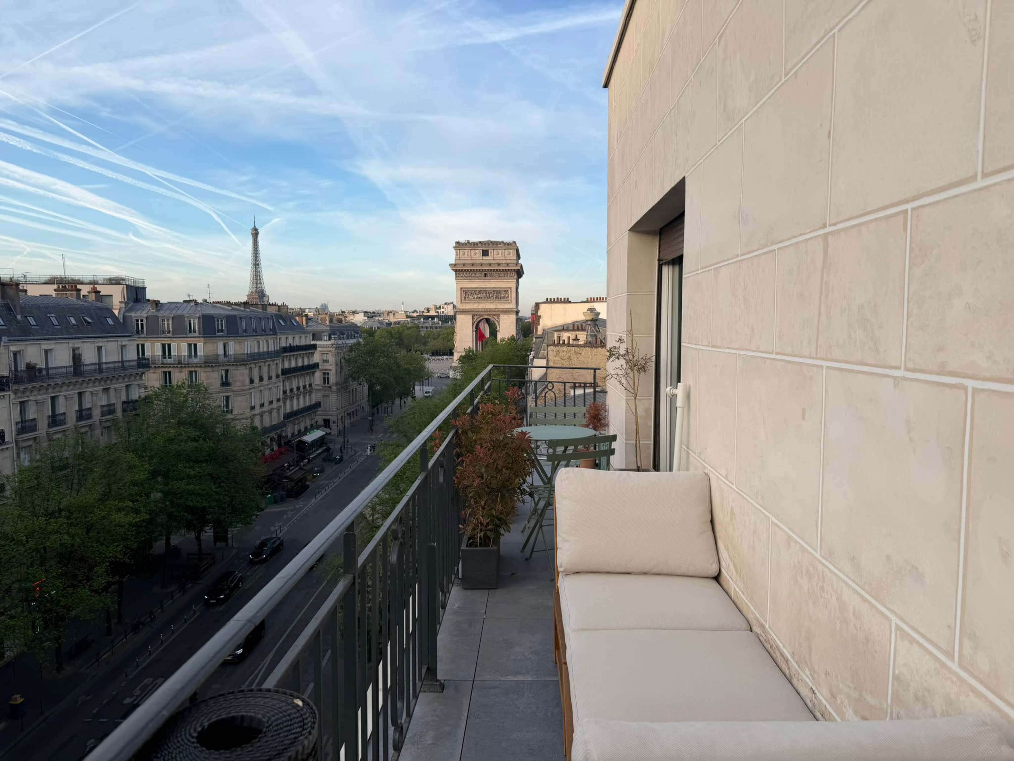 The image shows a balcony with a couch overlooking a Paris street, featuring the Arc de Triomphe and the Eiffel Tower in the distance.