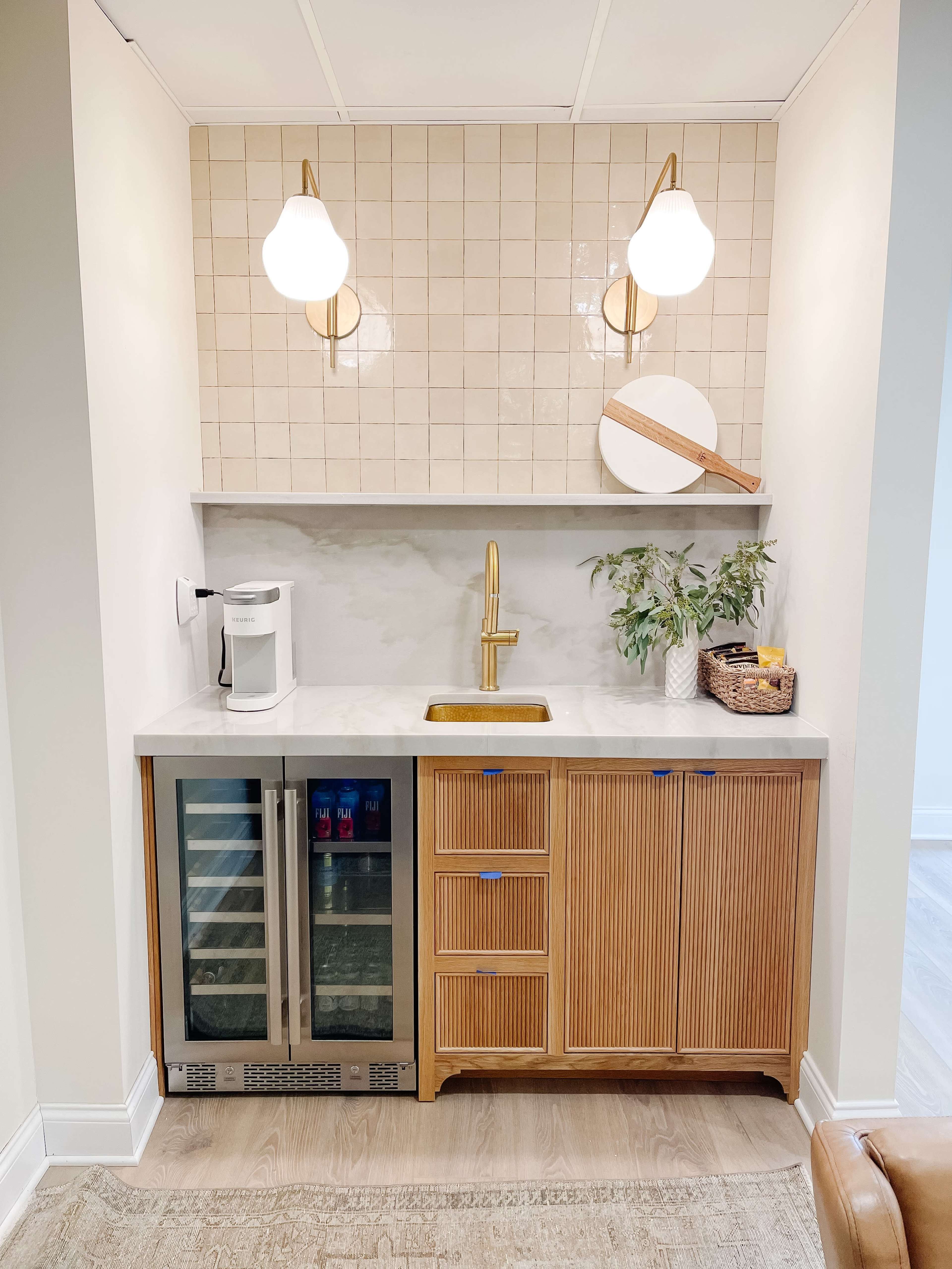 A modern kitchenette featuring a countertop sink, a mini fridge, wooden cabinetry with vertical slats, and decorative light fixtures above.