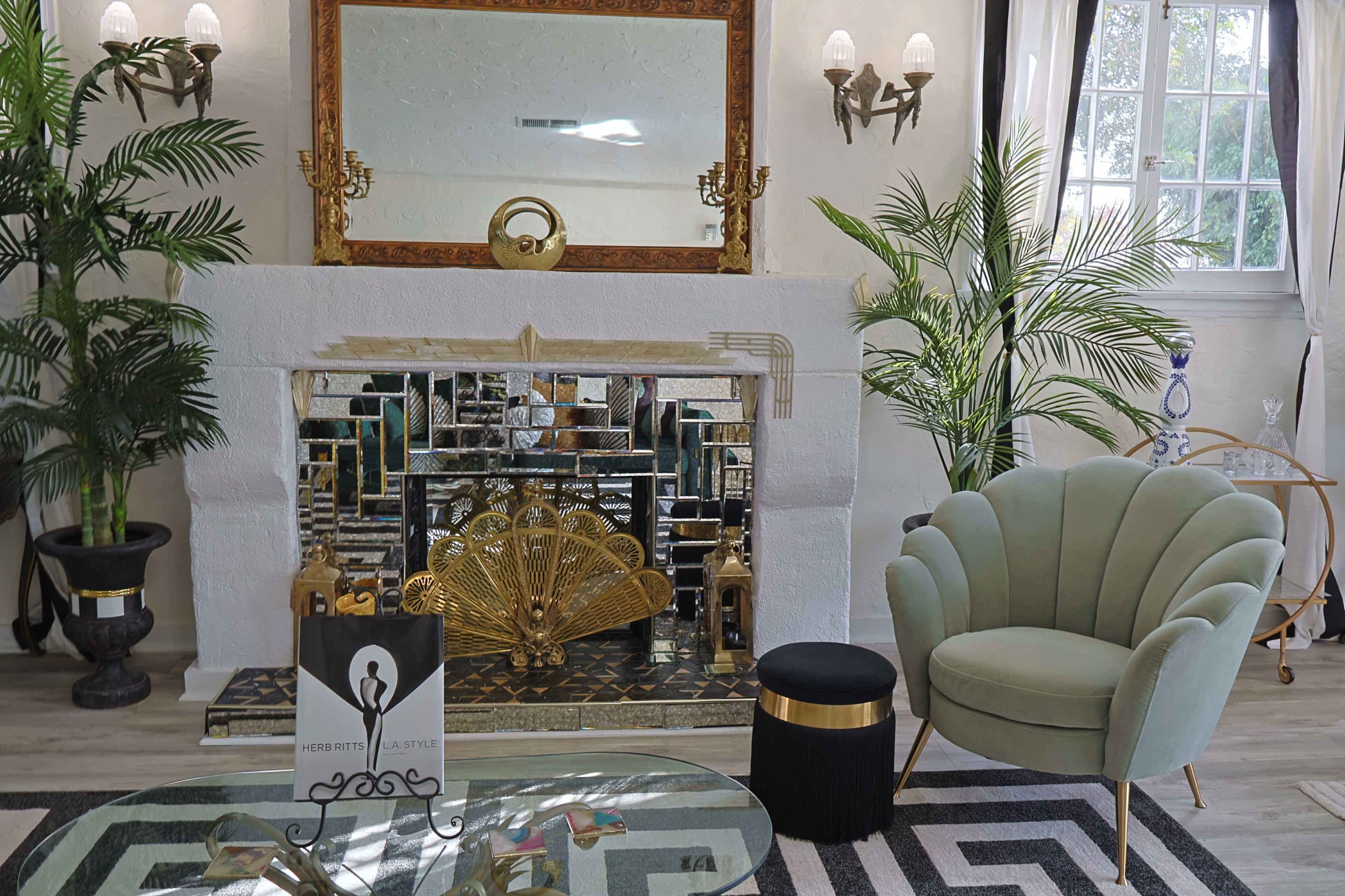 The image shows a modern living room with a decorative fireplace featuring a large mirror and palm plants, along with a green tufted chair and a small black side table.